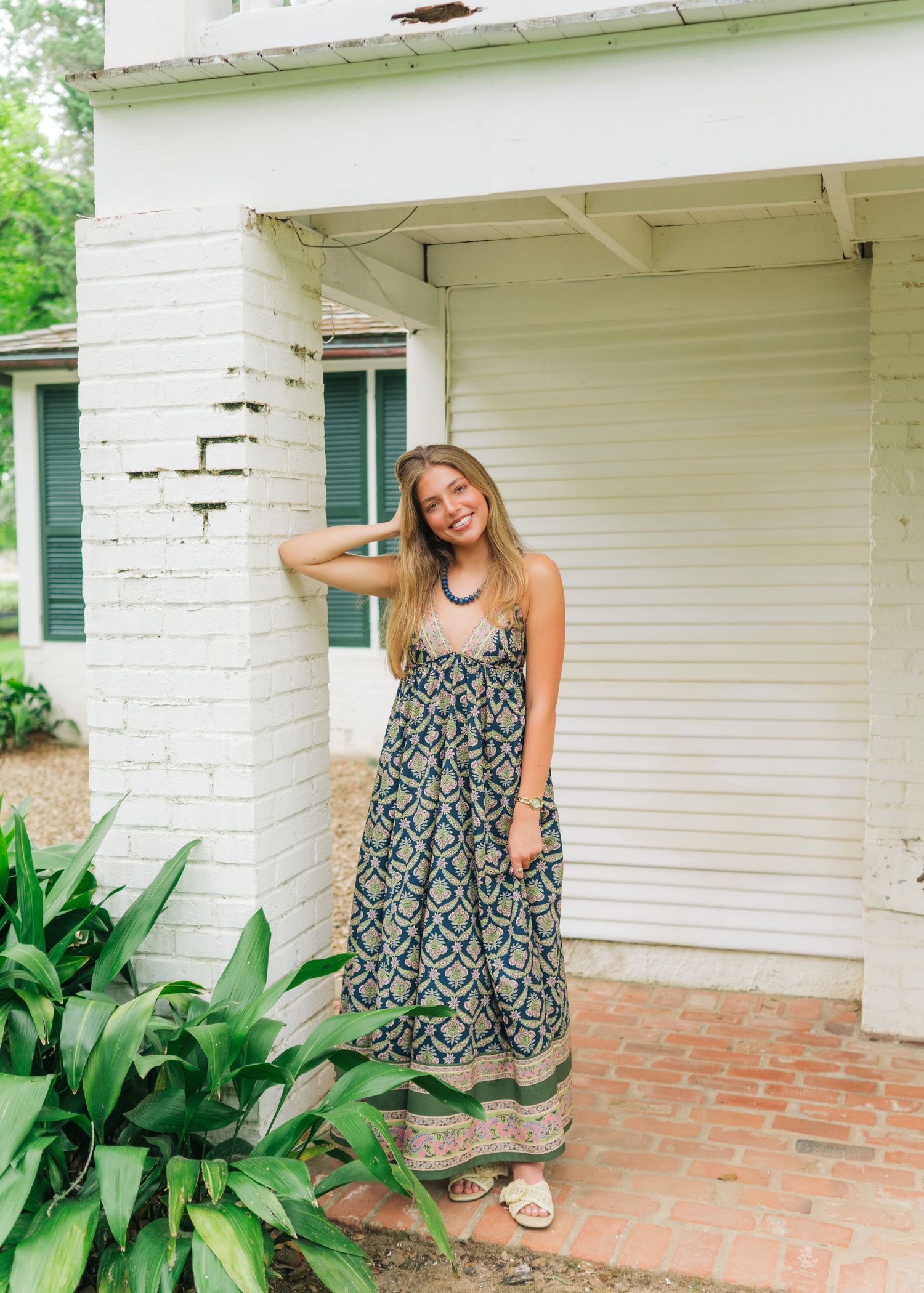 Young woman in a boho patterned maxi dress stands beside a white brick building with tropical plants nearby.