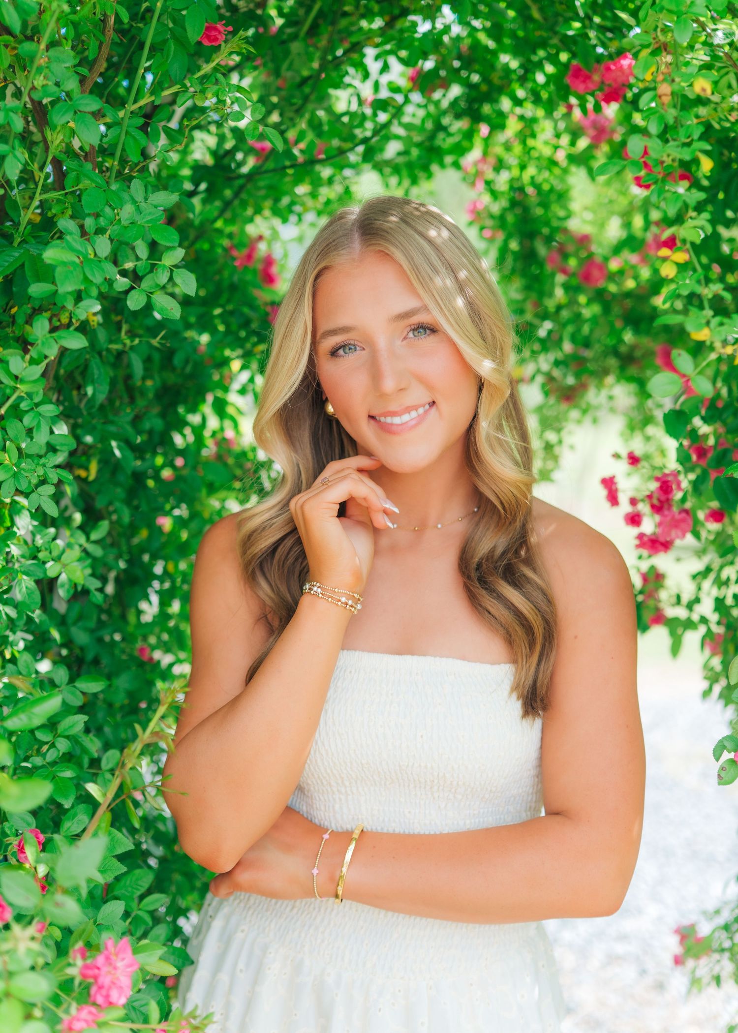 Smiling blonde woman in white dress posing among lush green and pink flowering vines.