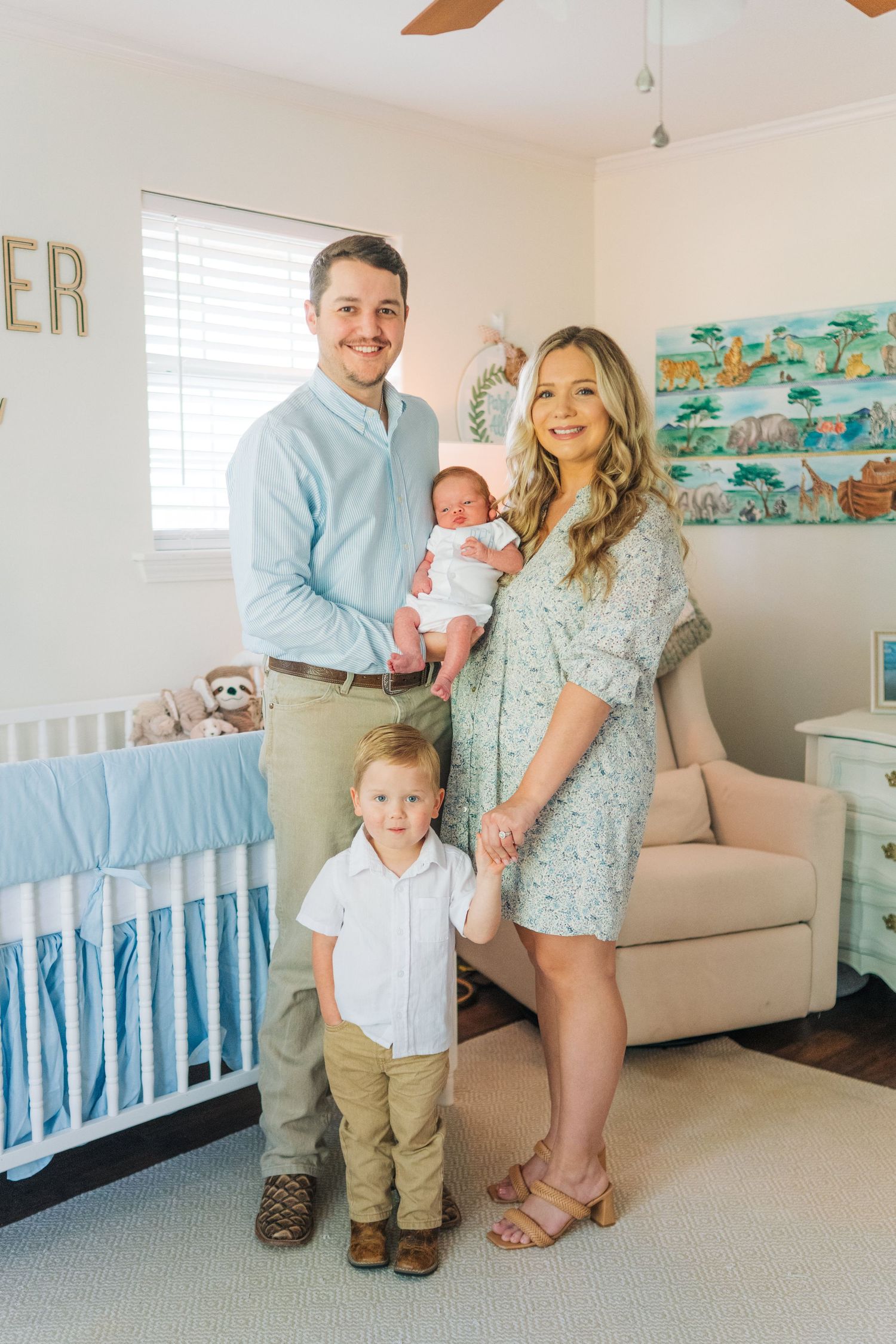Family of four poses in a safari-themed nursery, parents holding newborn and toddler beside a blue crib.