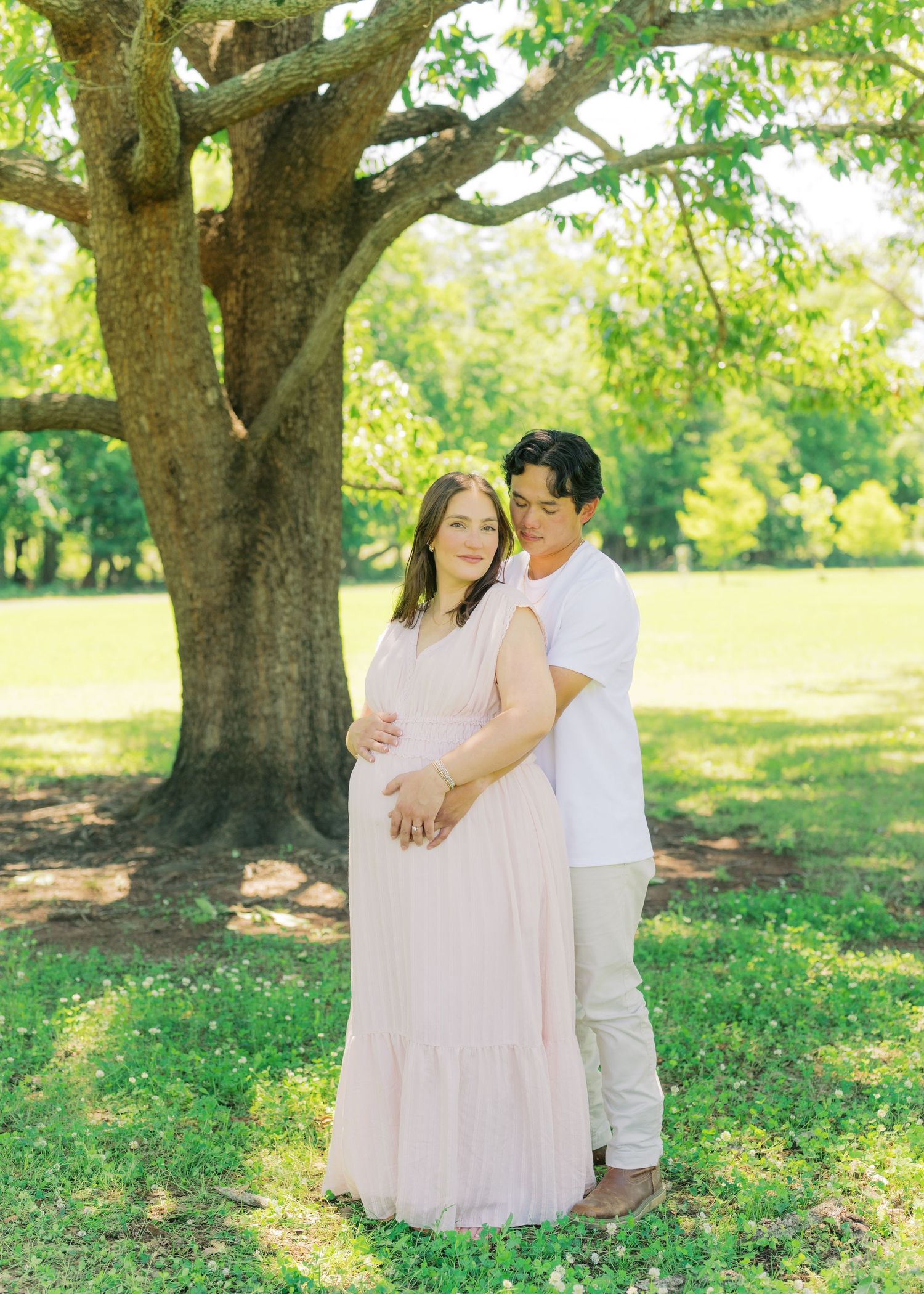 Couple sharing tender maternity portrait embrace under large tree in sunny green park.
