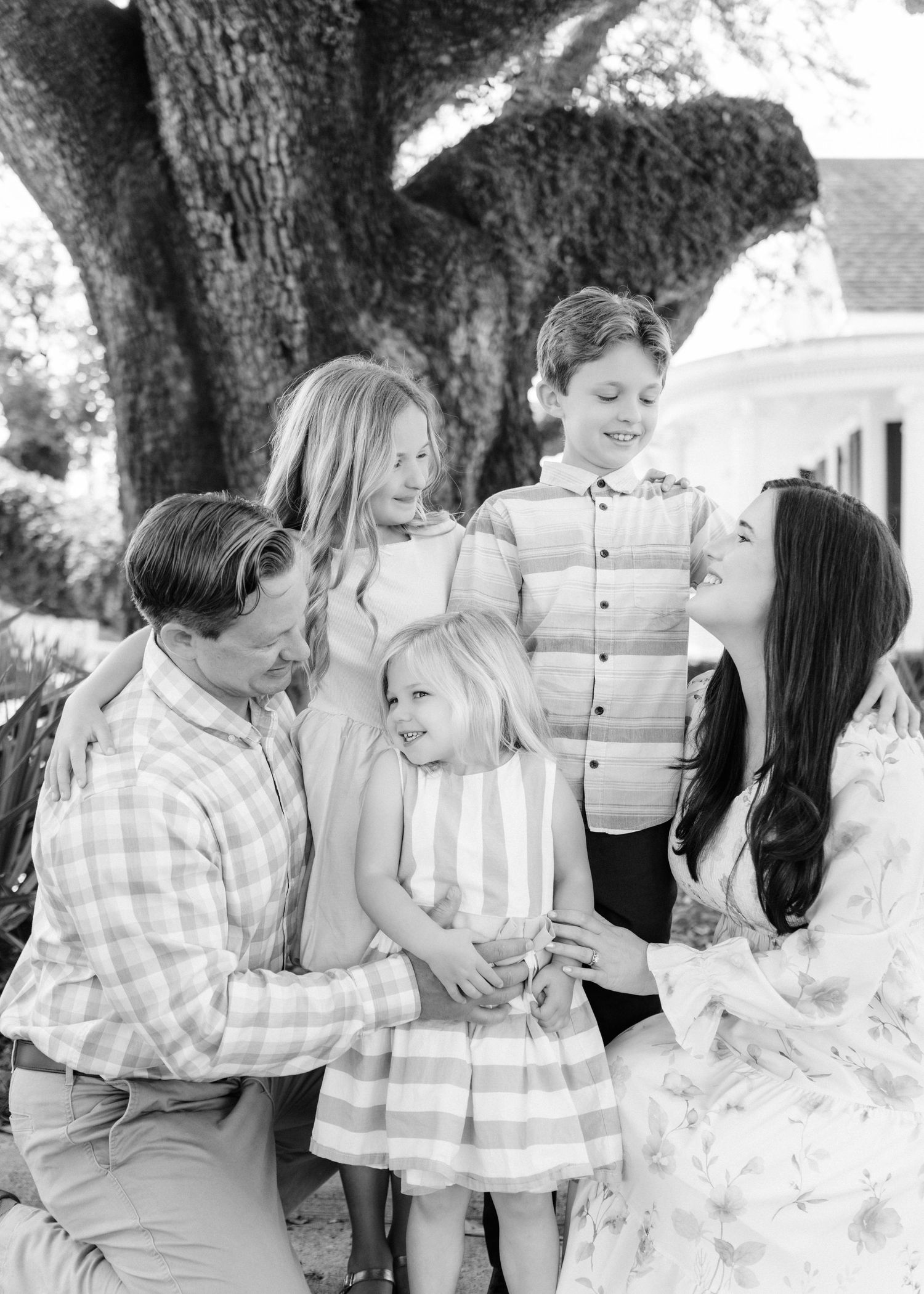 Black and white family portrait of parents with three young children smiling outdoors beneath a large oak tree.