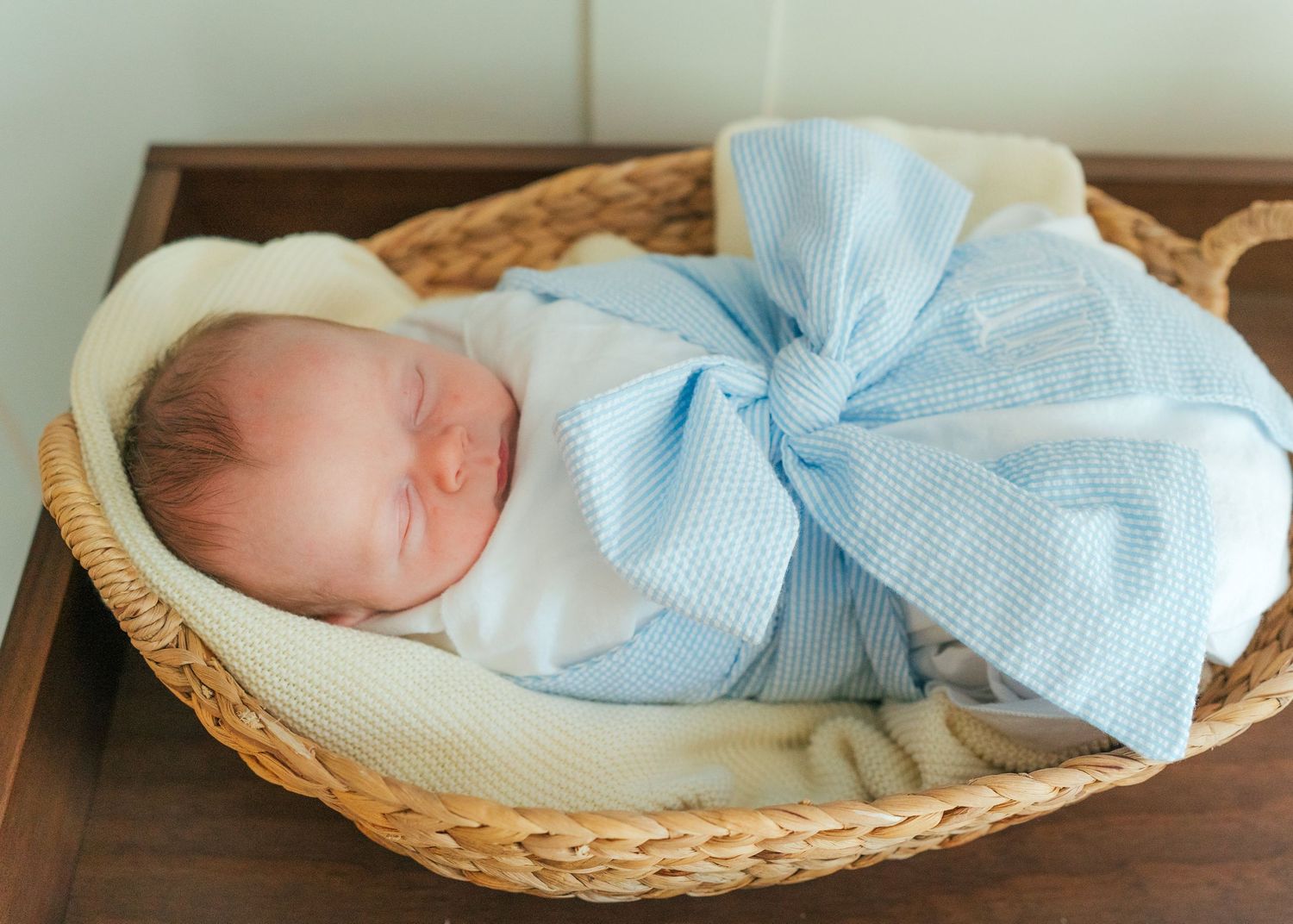 Newborn baby sleeping peacefully in a wicker basket wrapped in a white swaddle with a light blue bow.