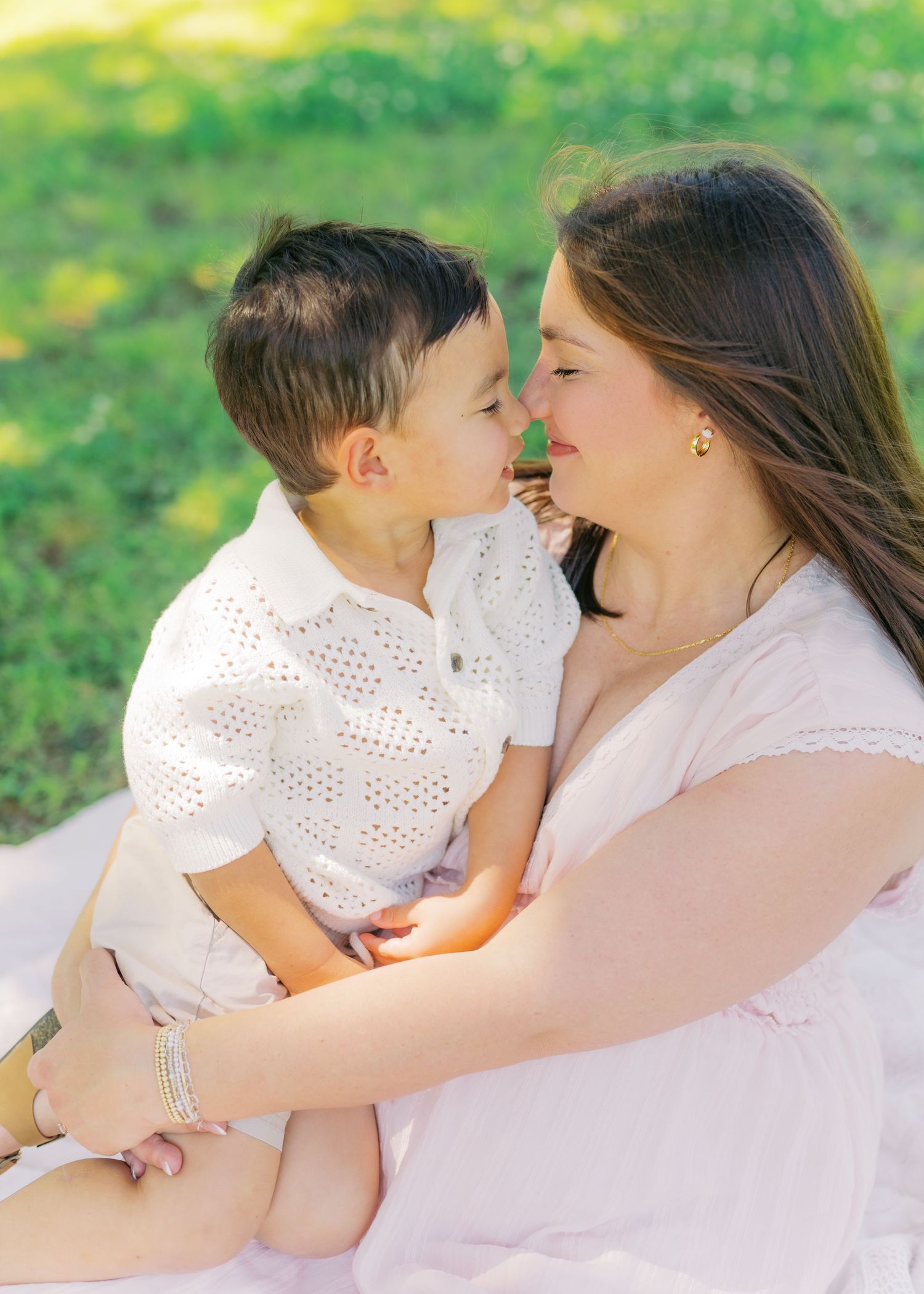 A mother and young boy share a sweet nose-to-nose moment while sitting on a blanket in a sunny green park.