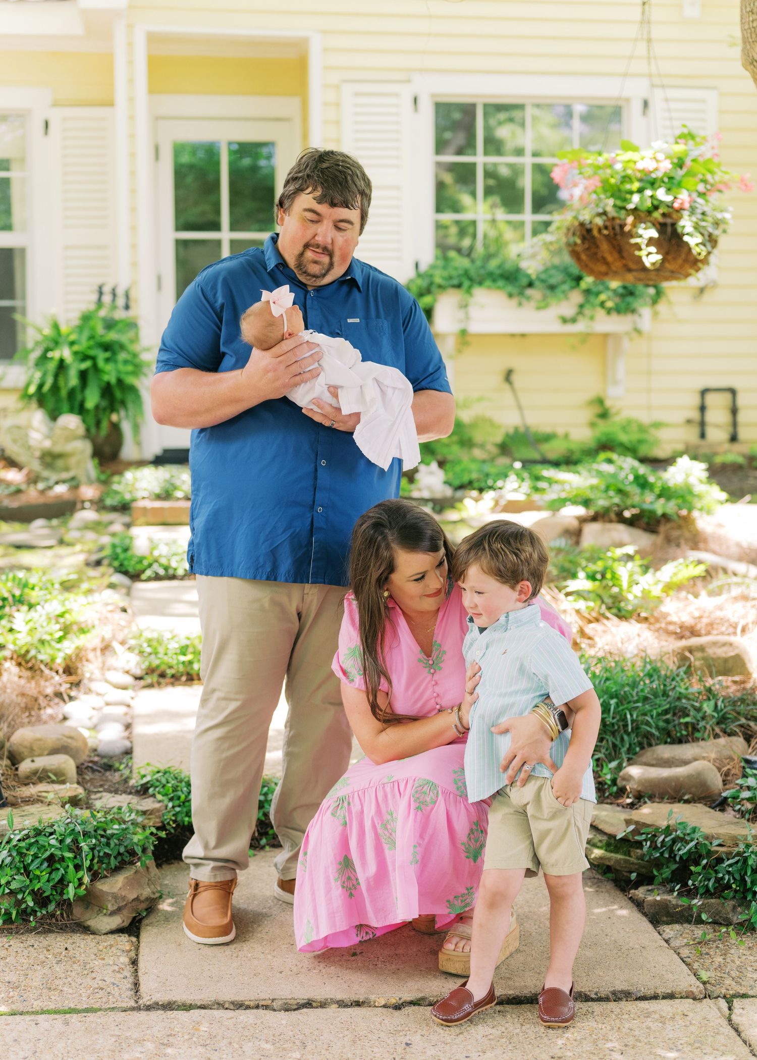 Family of four posing outdoors, father holding newborn, mother crouching with toddler son, beside a yellow house garden.