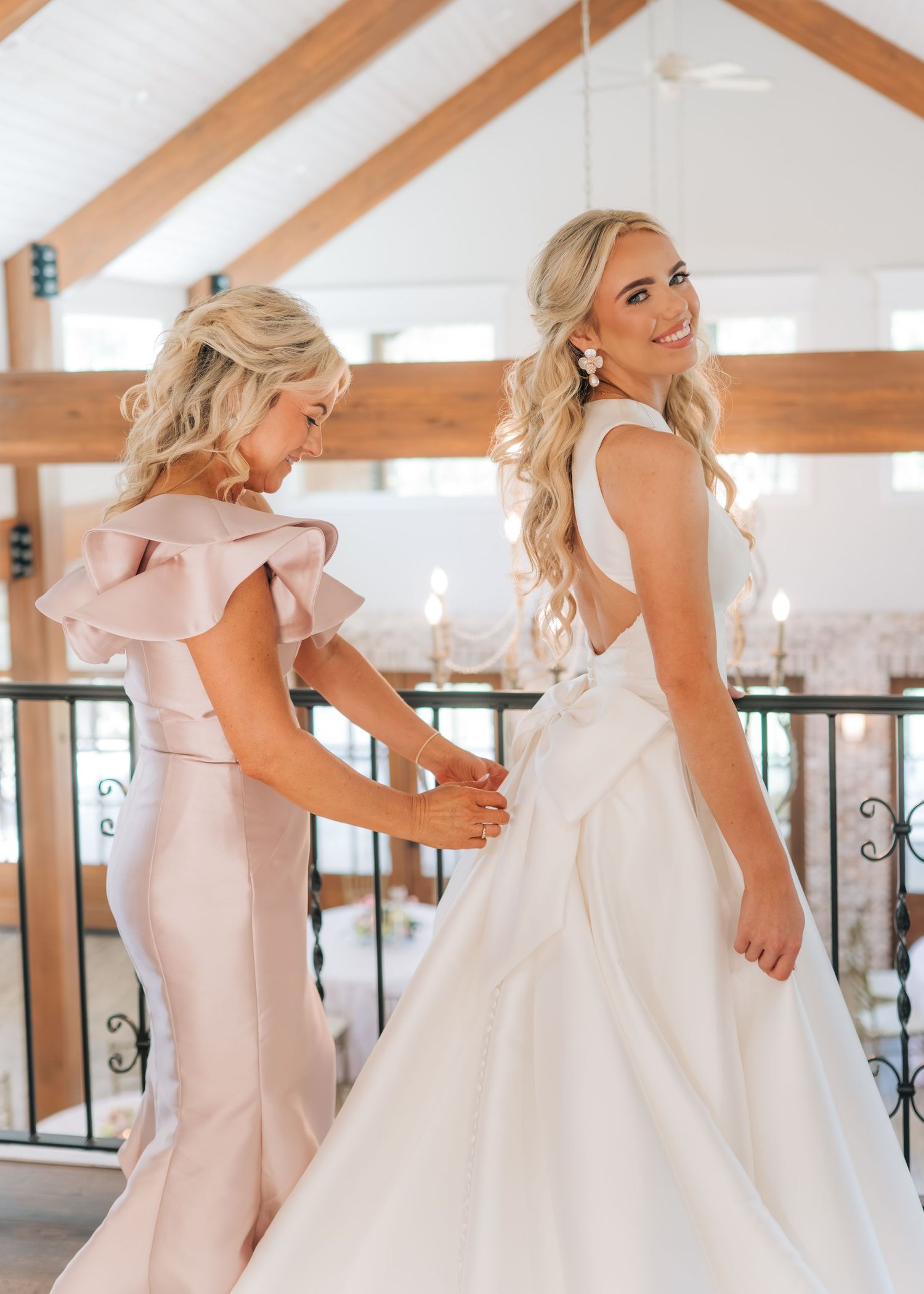 Bridesmaid in blush pink gown helping bride with white wedding dress in elegant venue.
