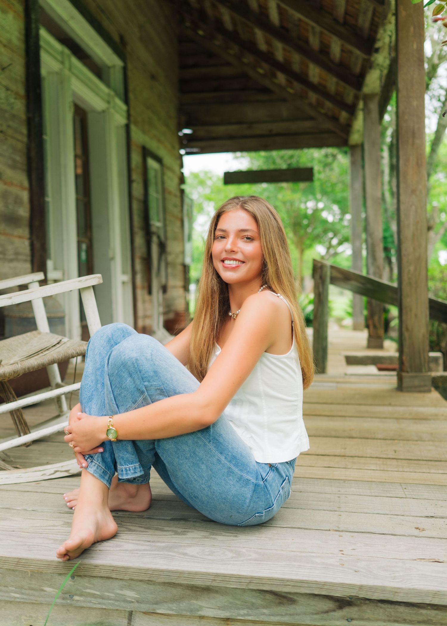 Young woman with long blonde hair sits smiling on a rustic wooden porch, wearing white tank top and blue jeans.