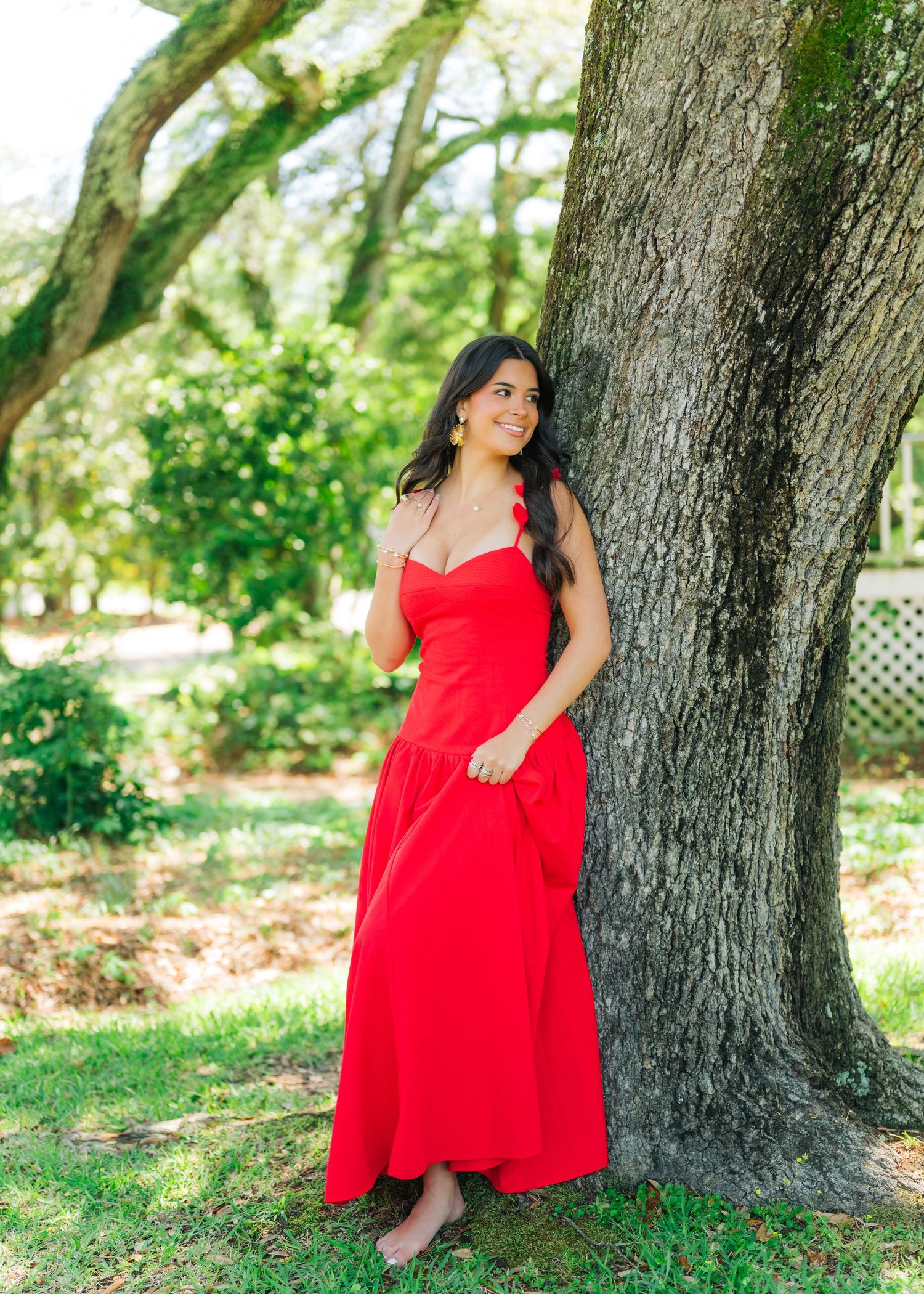 Young woman in elegant red maxi dress leans against large tree trunk in outdoor park setting.