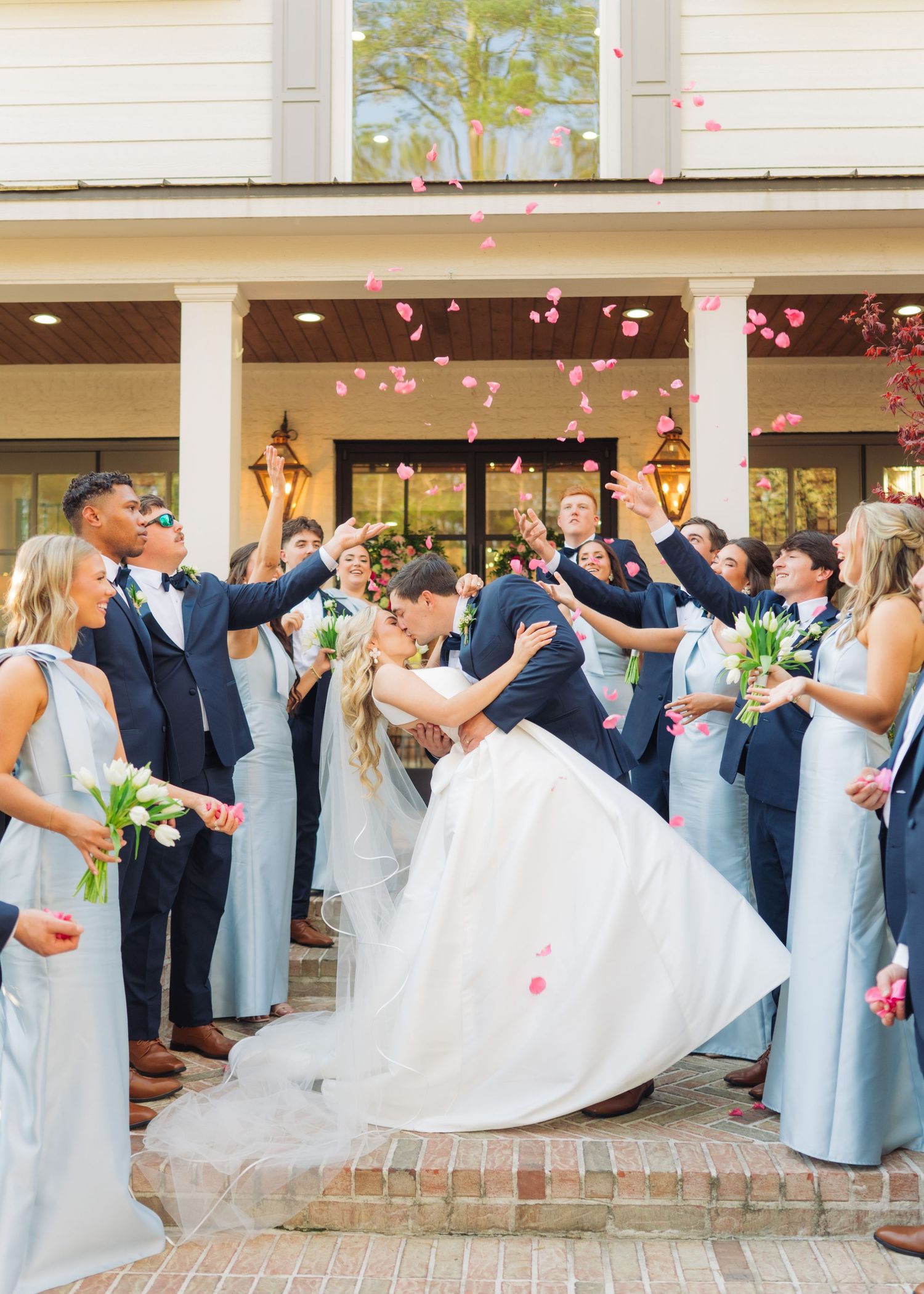 Bride and groom share a dip kiss surrounded by wedding party tossing pink rose petals outside elegant white venue.