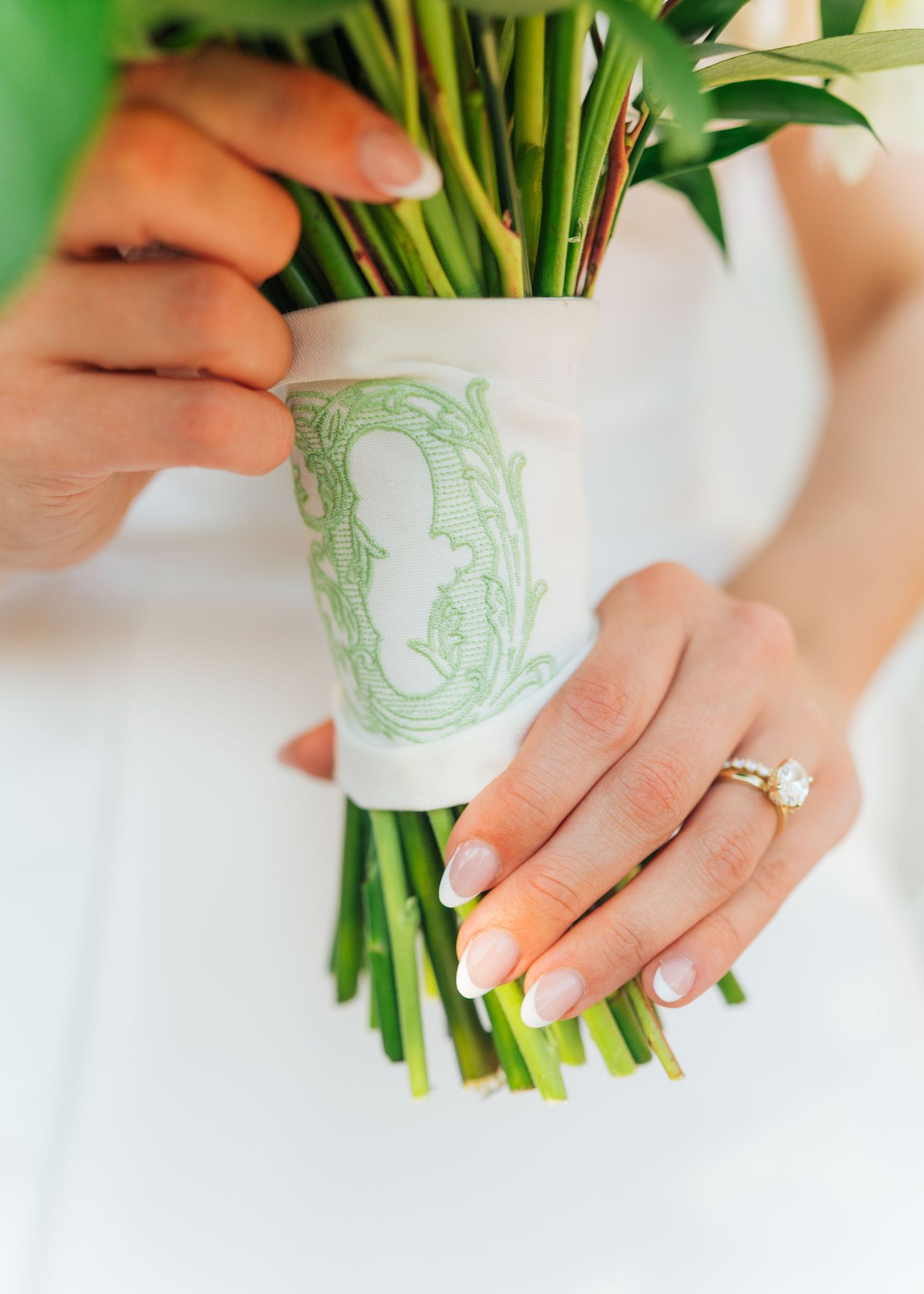 Close-up of bride holding bouquet wrapped in green monogram ribbon, showcasing diamond engagement ring and French manicure.
