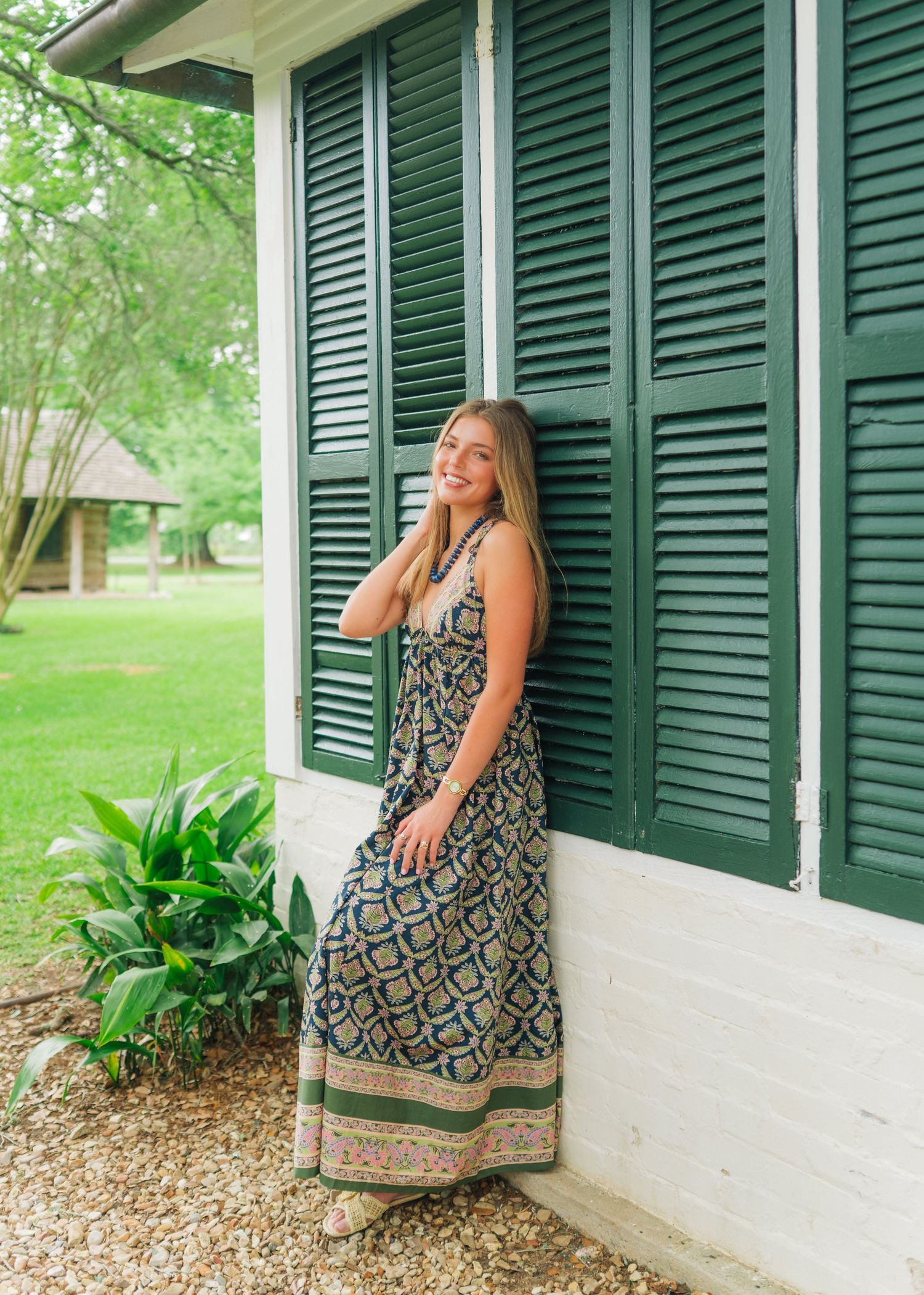 Woman in patterned maxi dress smiling beside green shuttered window on porch.