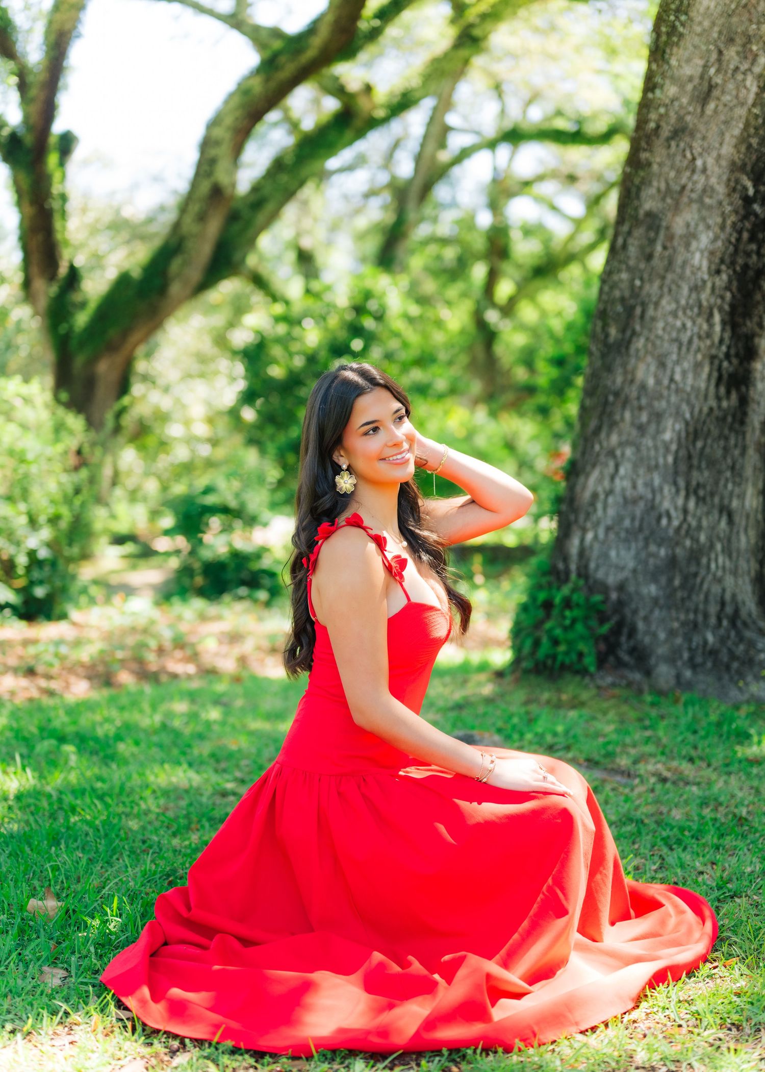 Young woman in flowing red gown sitting gracefully on grass beneath large oak tree.