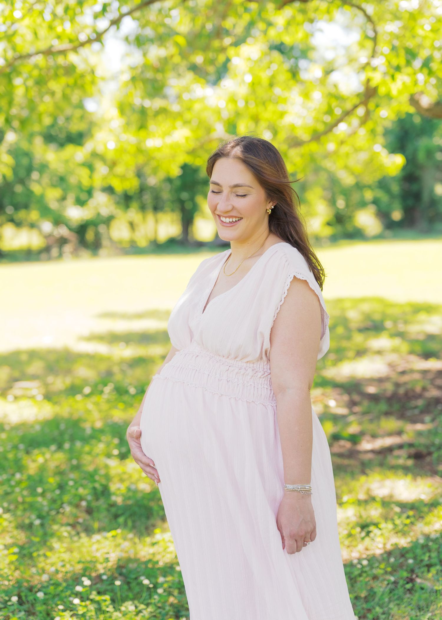 Smiling pregnant woman in blush pink dress cradling baby bump outdoors in lush green park.