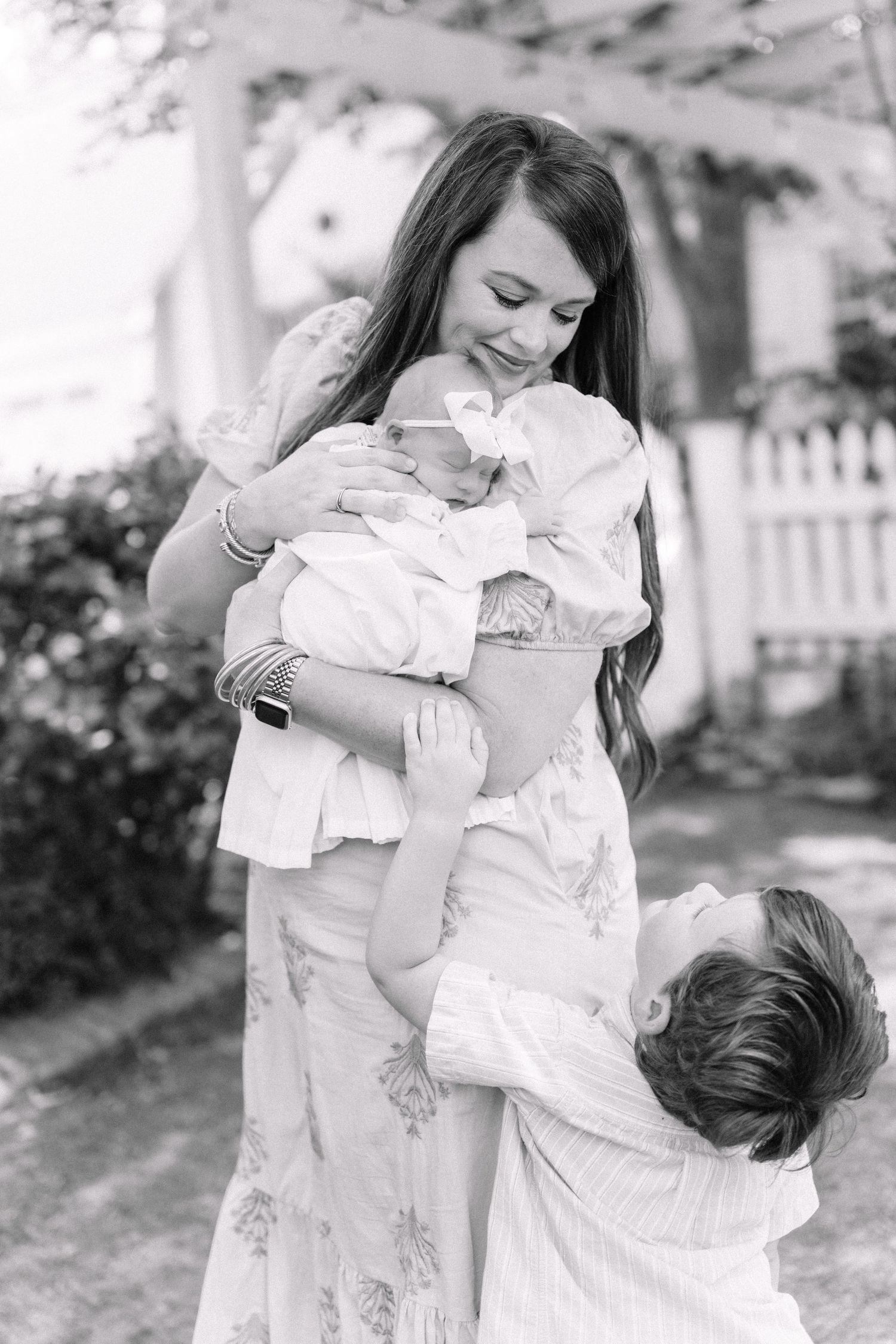 Black and white photo of a smiling mother holding newborn baby while older child looks up adoringly.