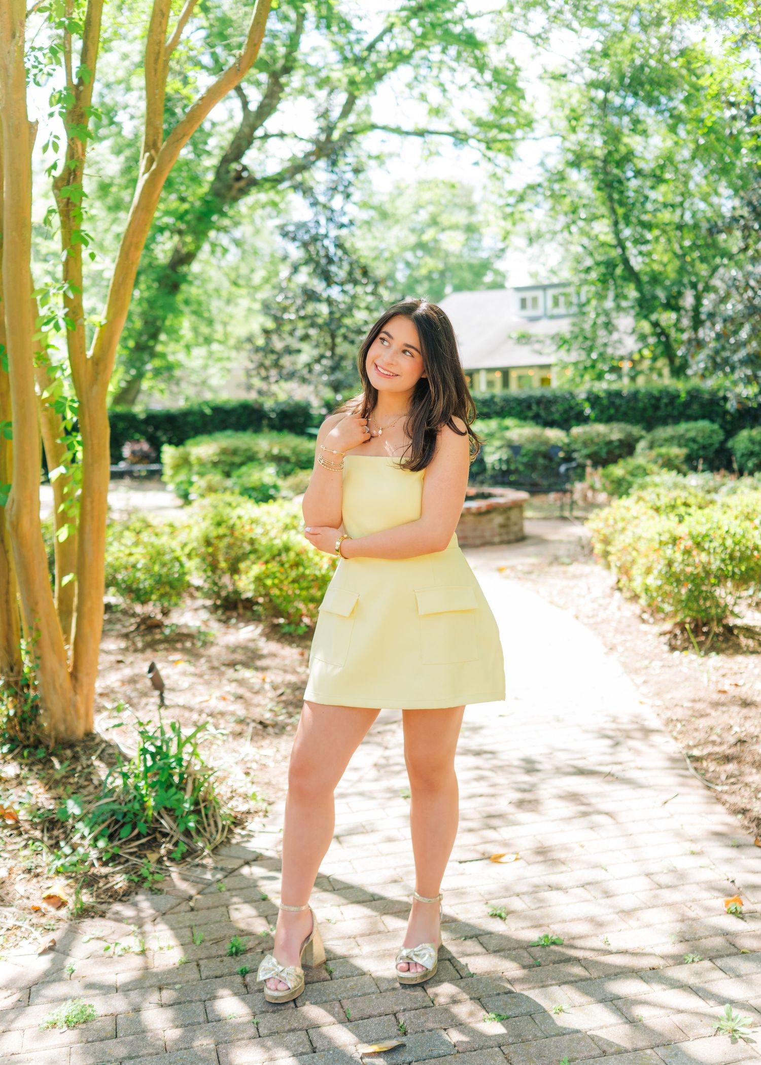 Young woman in yellow strapless mini dress posing on brick path in lush garden with green hedges.