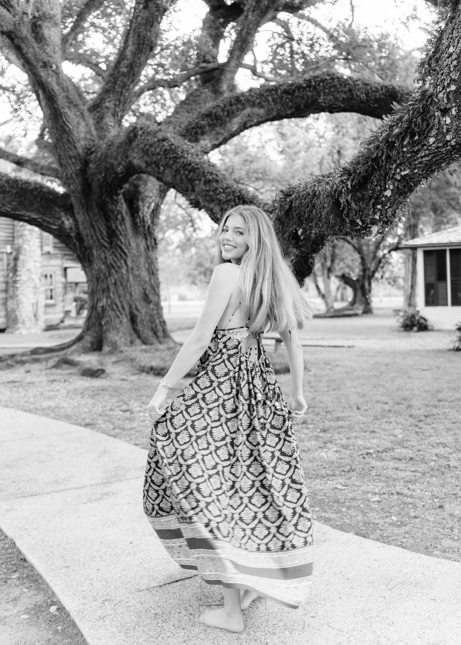 Black and white photo of woman twirling in patterned maxi dress under large oak tree.