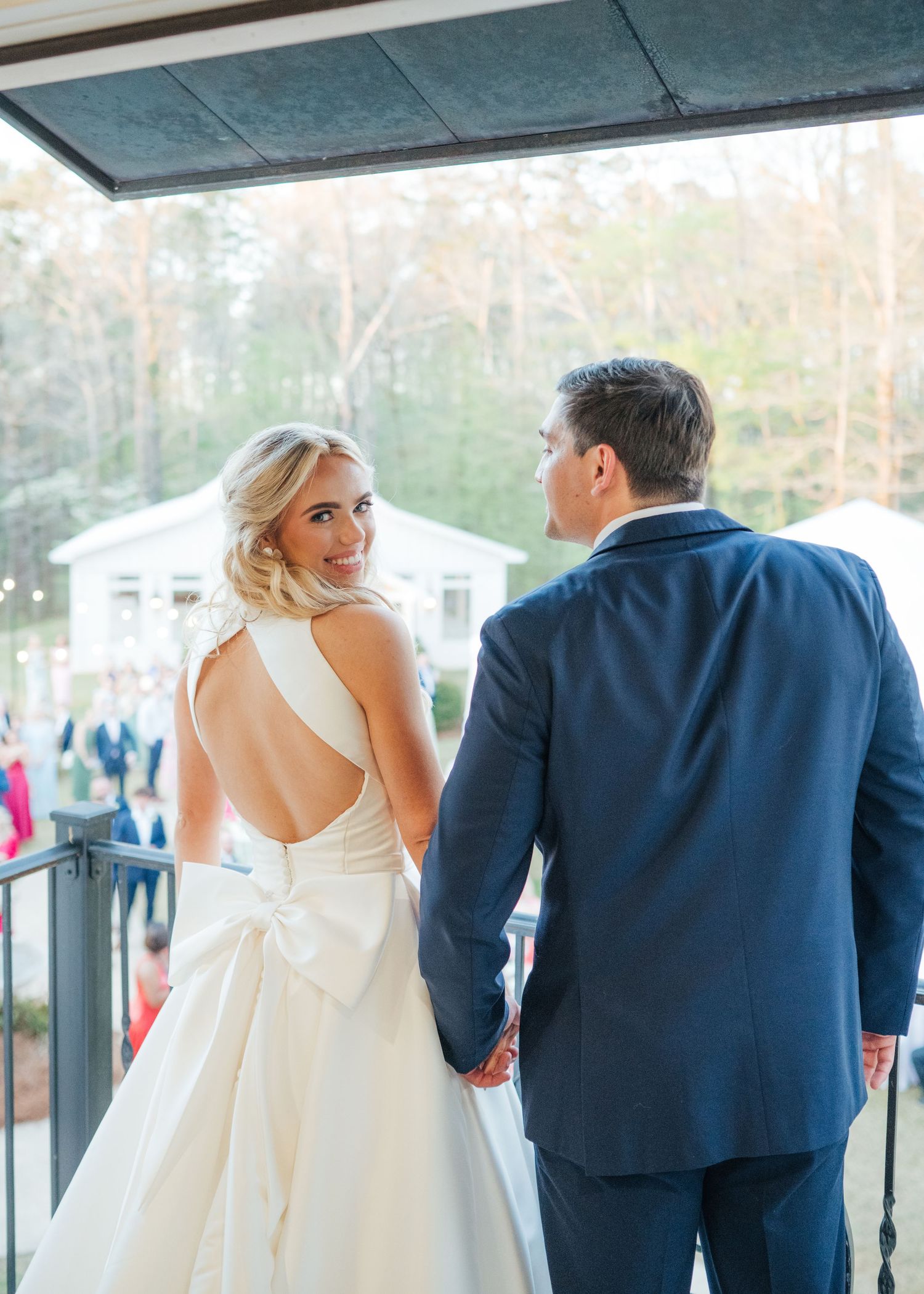 Bride in backless gown with bow holds groom's hand on balcony overlooking outdoor wedding guests.