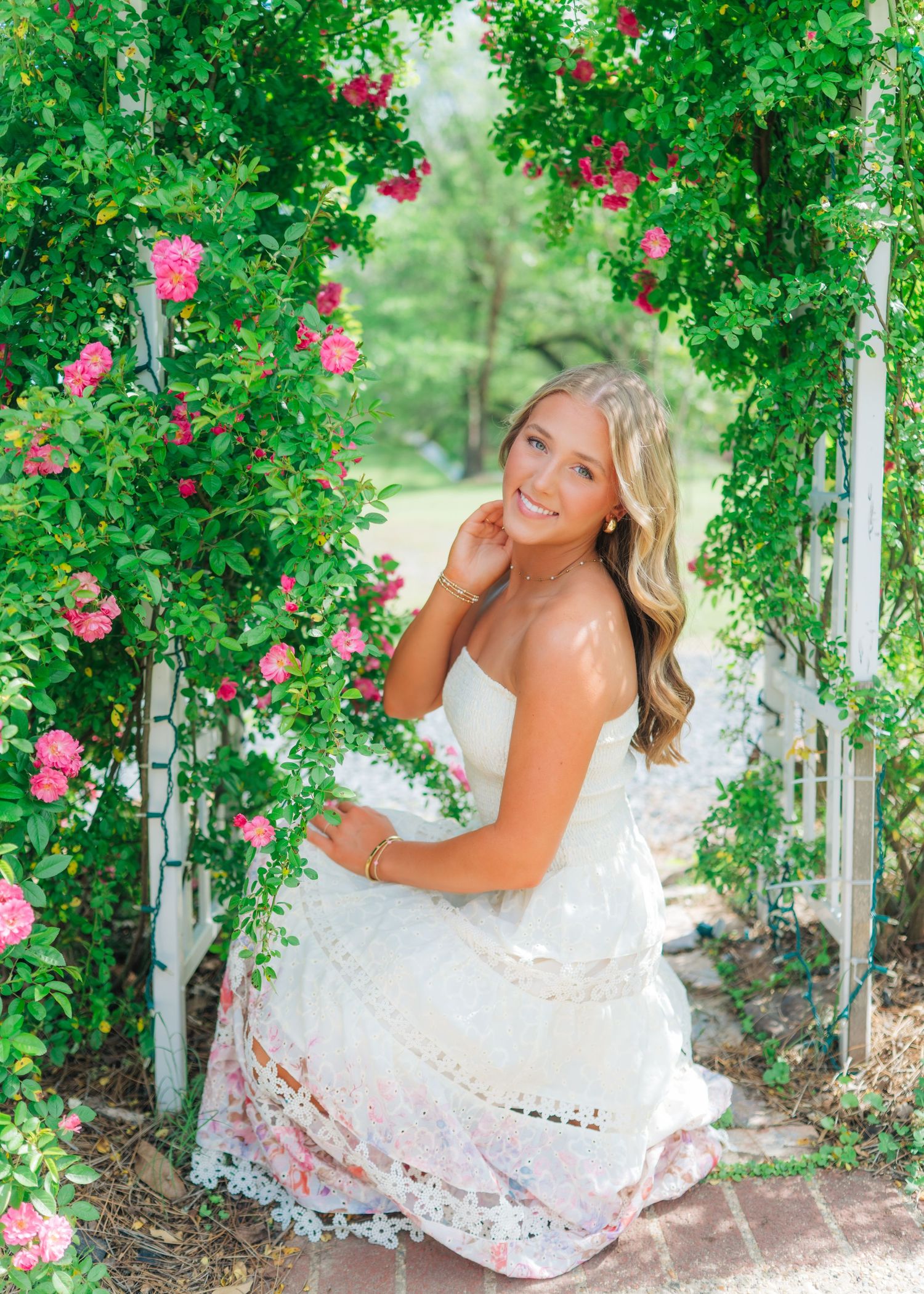Blonde teen in white floral lace dress sits beneath pink rose archway for elegant senior portrait.