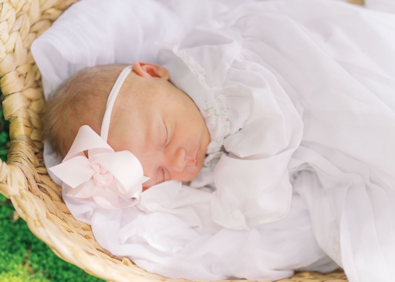 Newborn baby girl sleeping peacefully in a wicker basket, wrapped in white fabric with a pink bow headband.