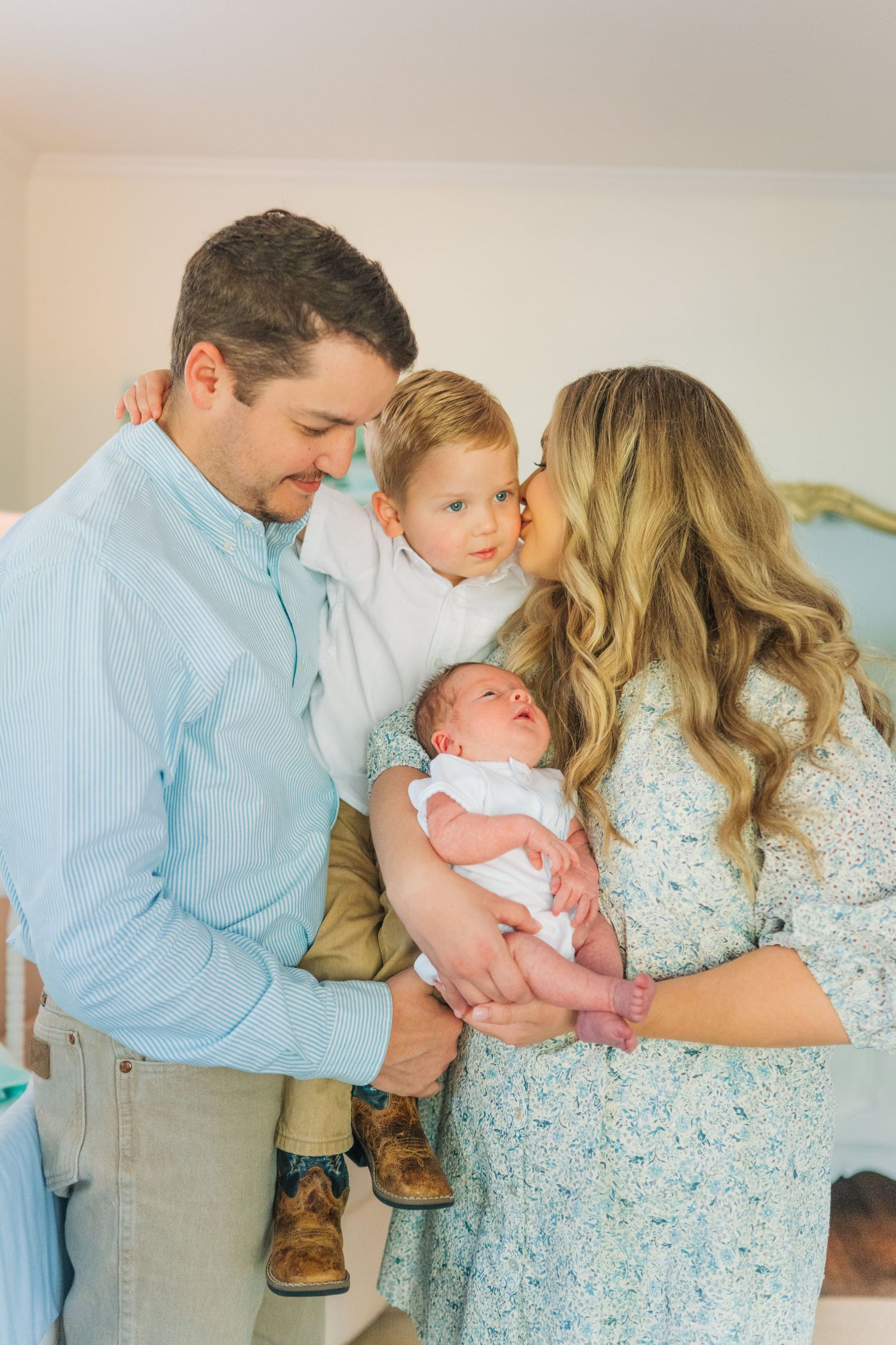 A young family of four poses indoors, parents holding a newborn and toddler in coordinating blue and floral outfits.
