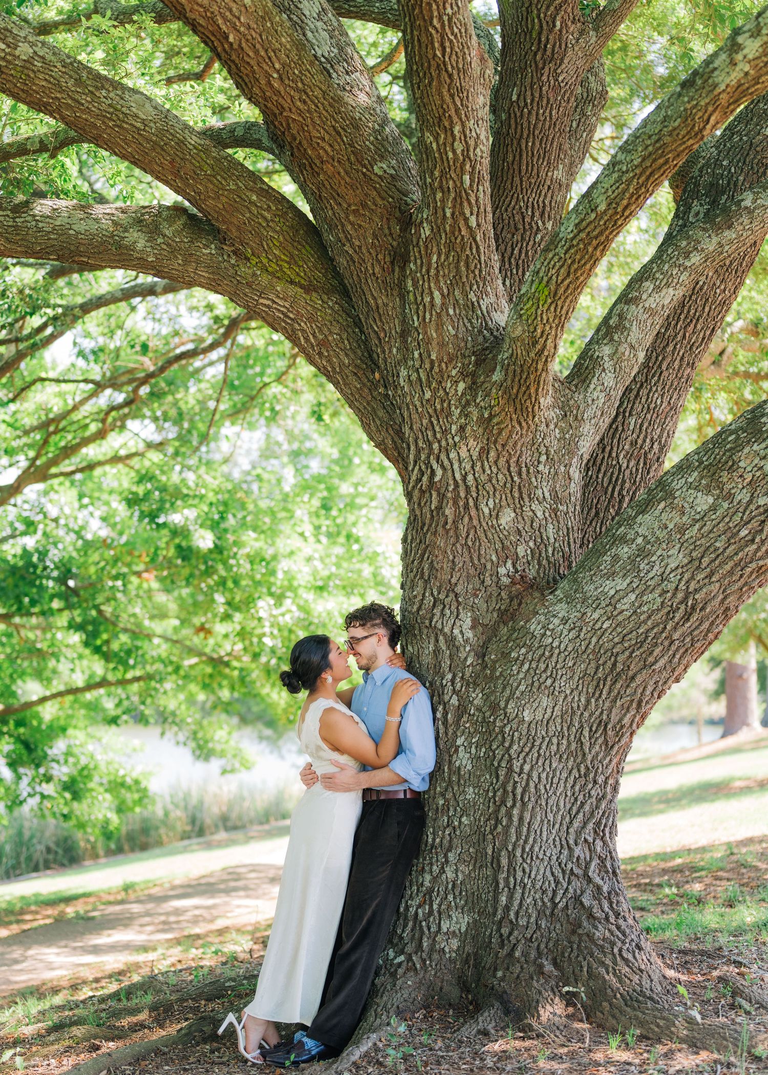 Couple sharing a romantic moment leaning against a large oak tree in a lush green park setting.