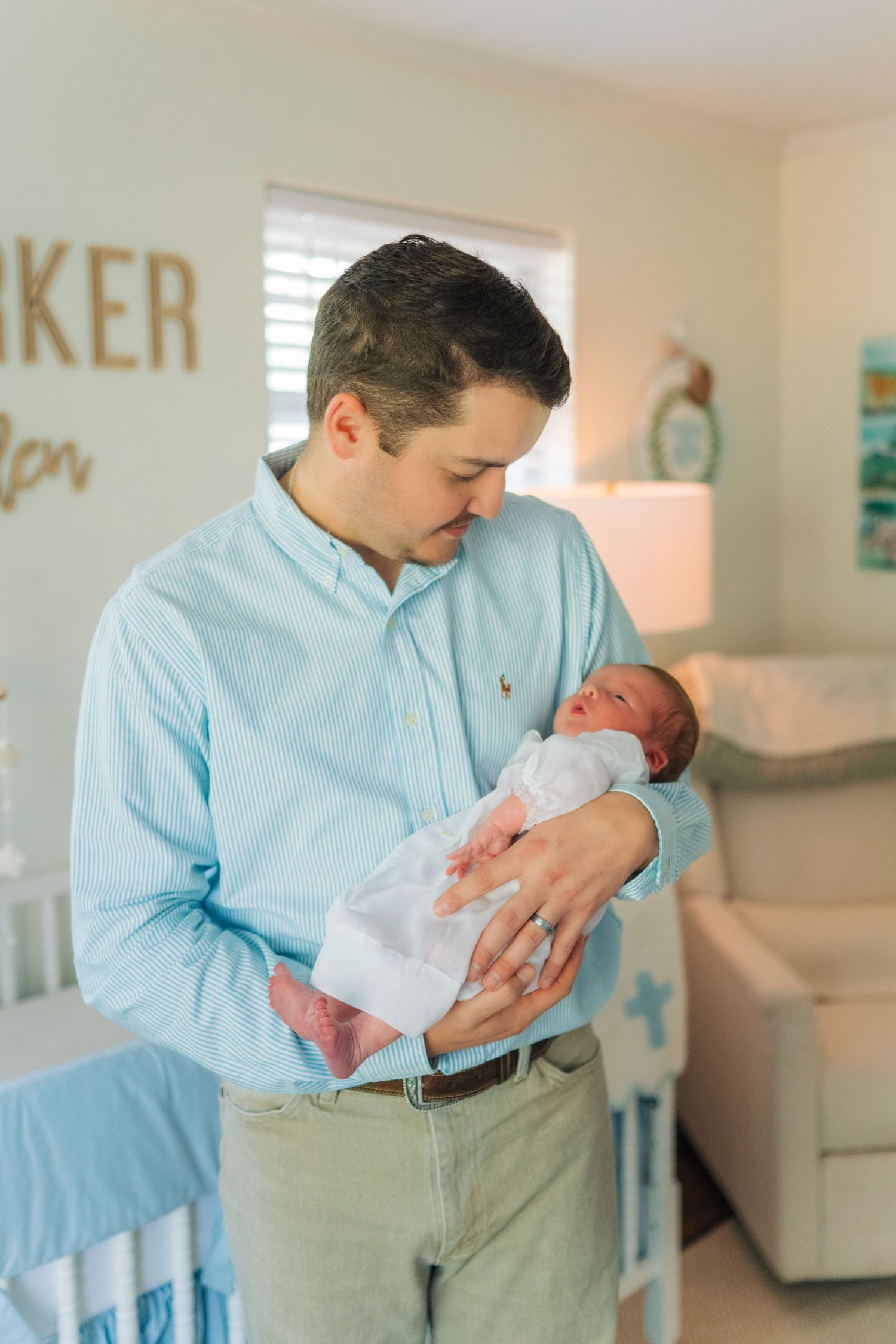 Father in light blue shirt tenderly holds swaddled newborn baby in a soft, decorated nursery room.