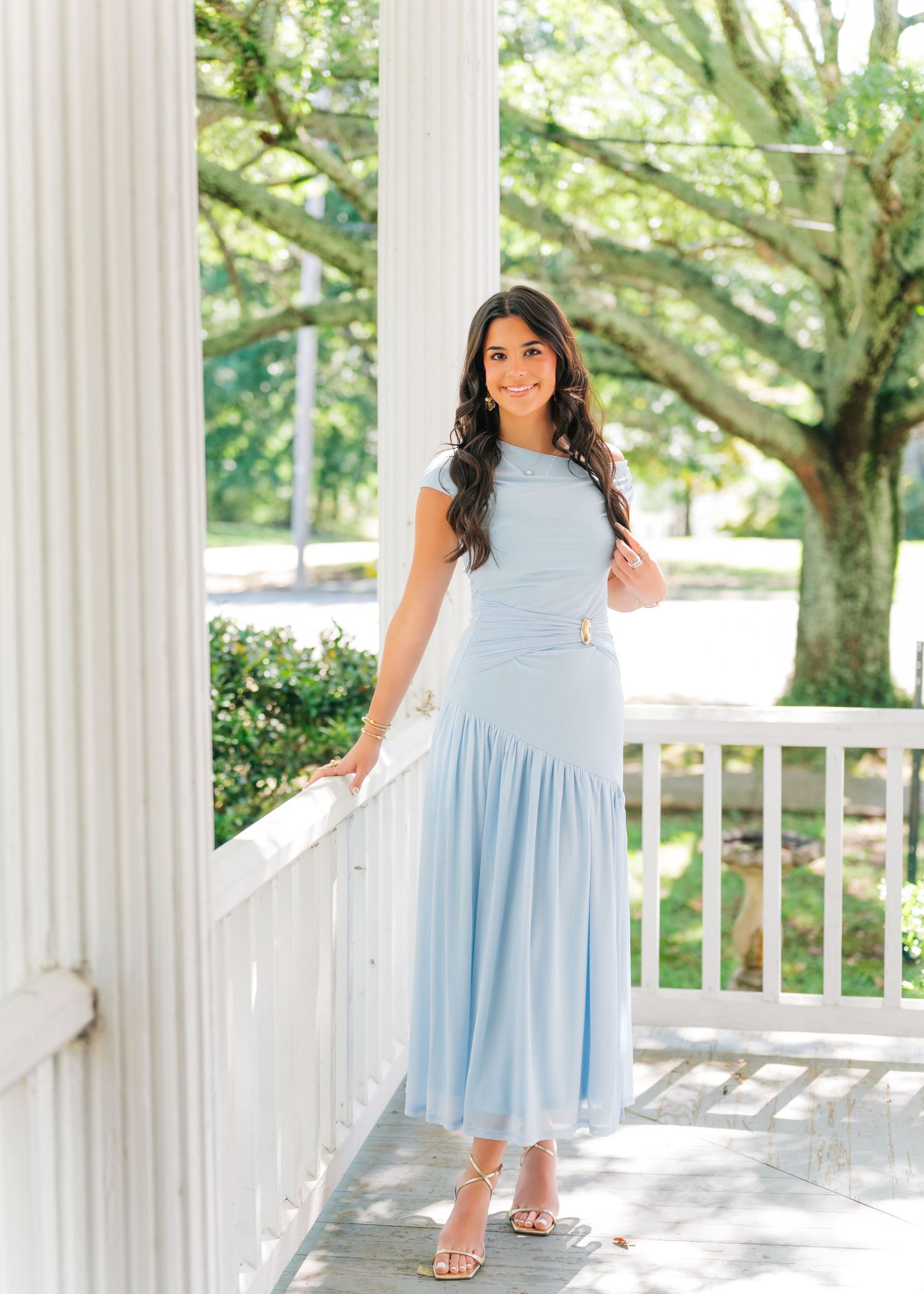 Woman in light blue midi dress posing on white porch with columns and green trees in background.
