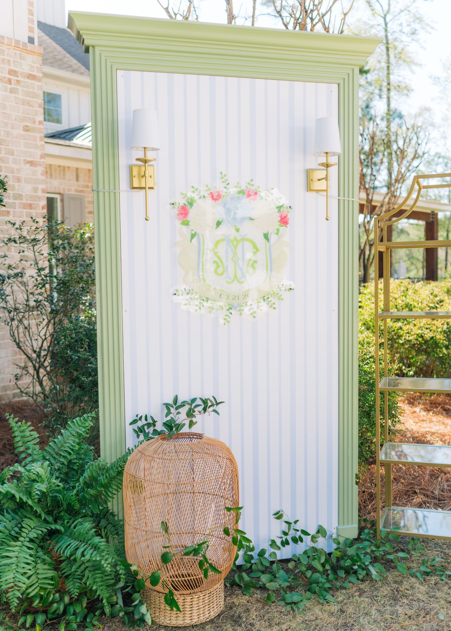 Elegant outdoor wedding monogram backdrop with green frame, floral crest, and wicker chair.