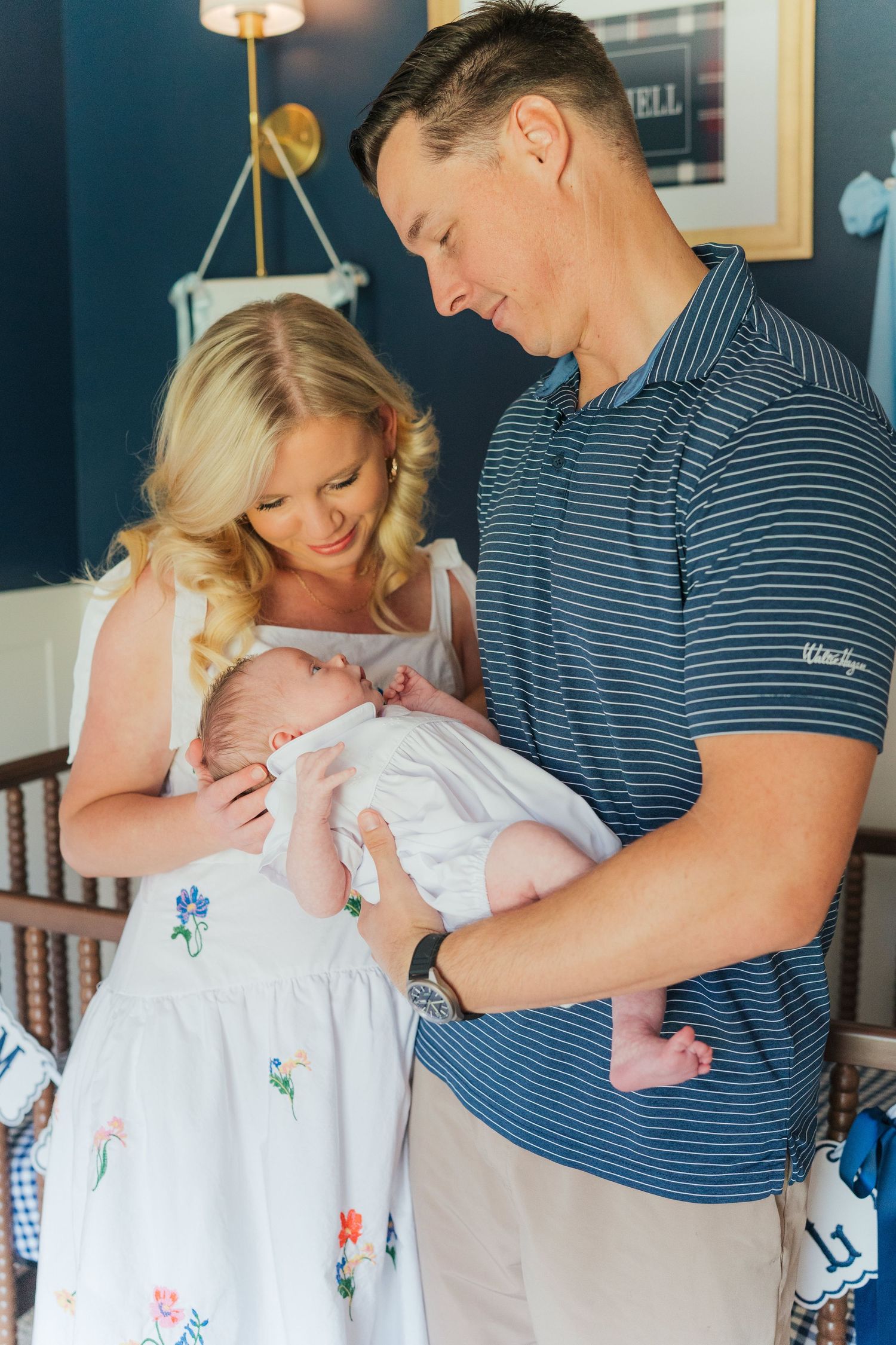 New parents lovingly gaze at their newborn baby in a navy blue nursery, mother in floral dress, father in striped polo.