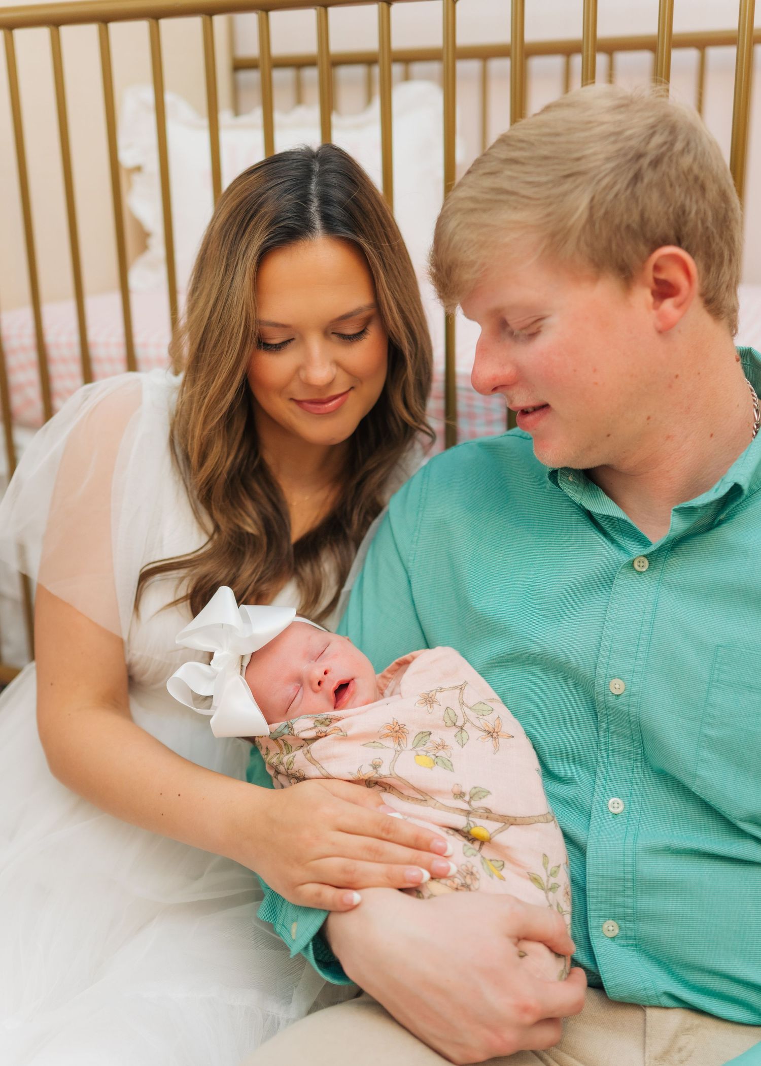 New parents in teal and white hold swaddled newborn baby with bow in nursery.