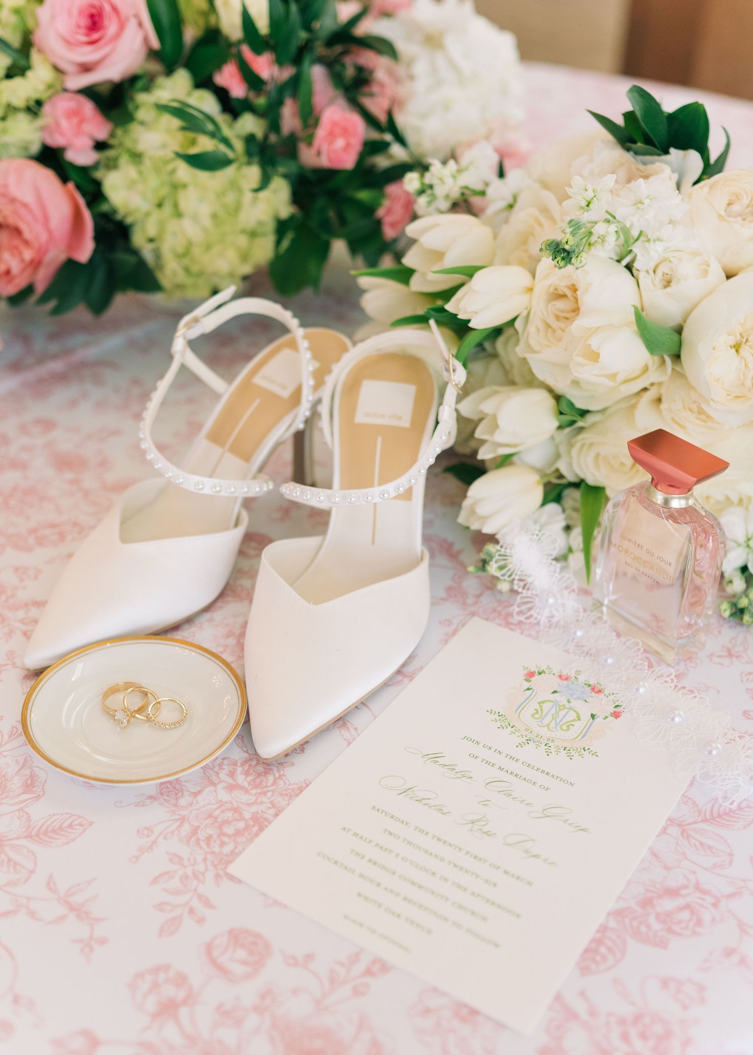White bridal heels with ankle straps beside wedding rings, floral bouquet, perfume, and elegant invitation on pink tablecloth.