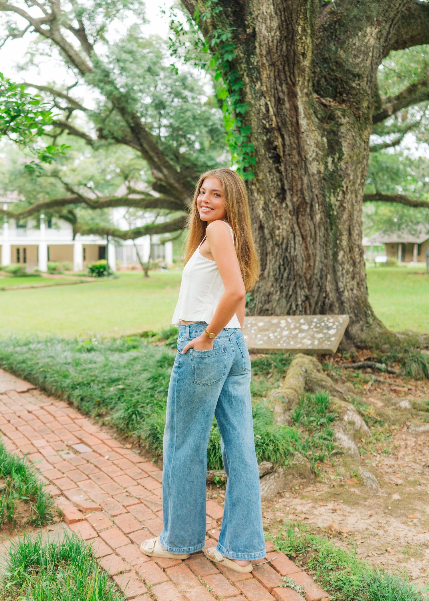 Young woman in wide-leg jeans and white top poses on brick path beside large oak tree.