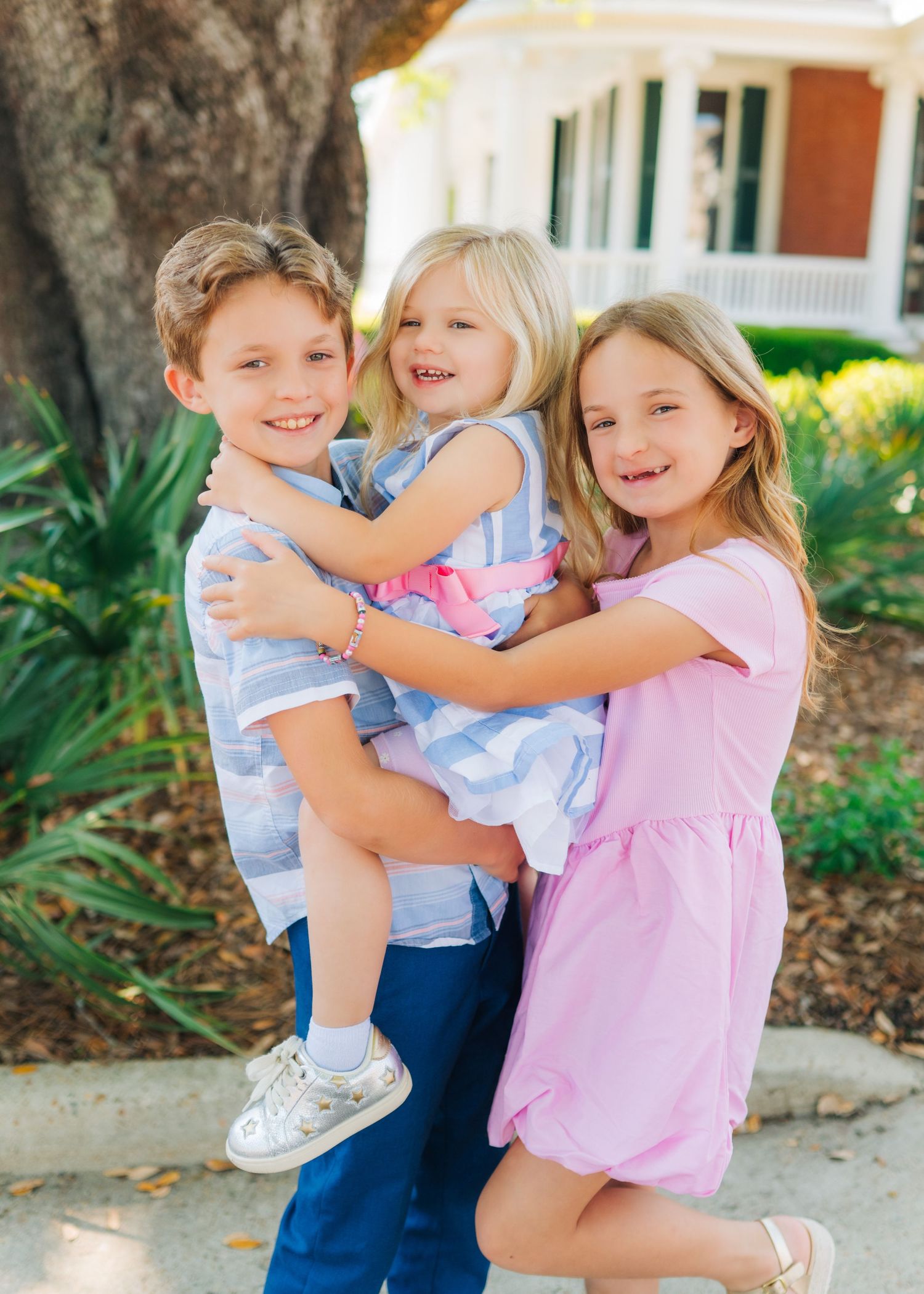 Three smiling children hugging outdoors near a tree, wearing pastel outfits, with a white house in the background.