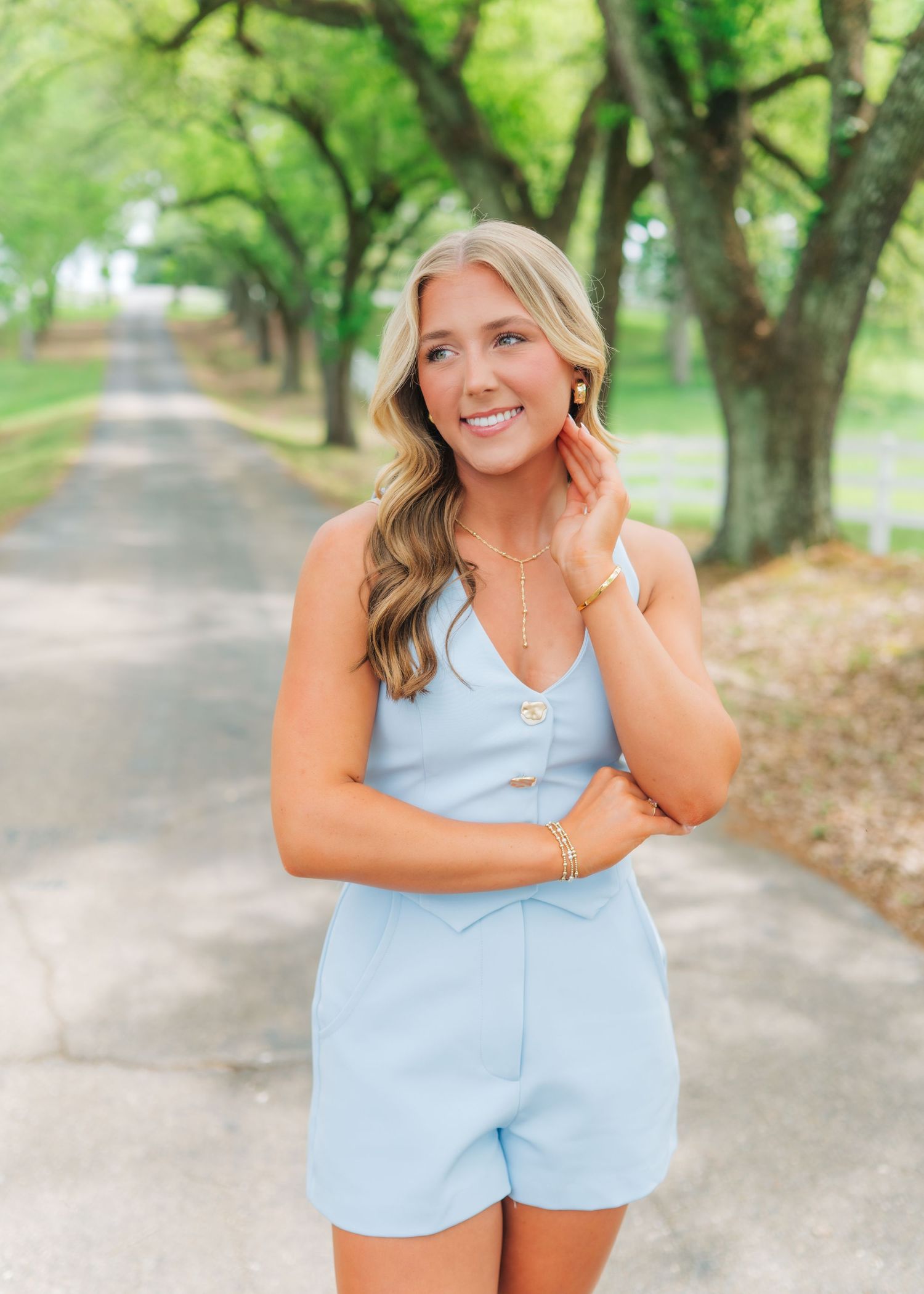 Young woman in light blue romper smiling on a tree-lined path, wearing gold jewelry on a sunny day.