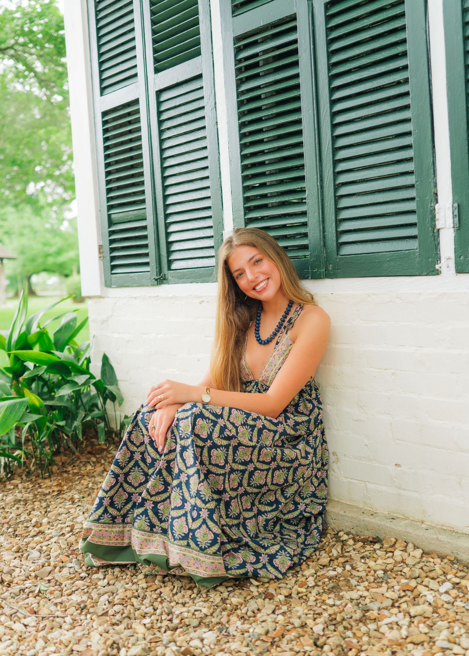 Smiling young woman in patterned maxi dress sits on gravel beside green shuttered white building.