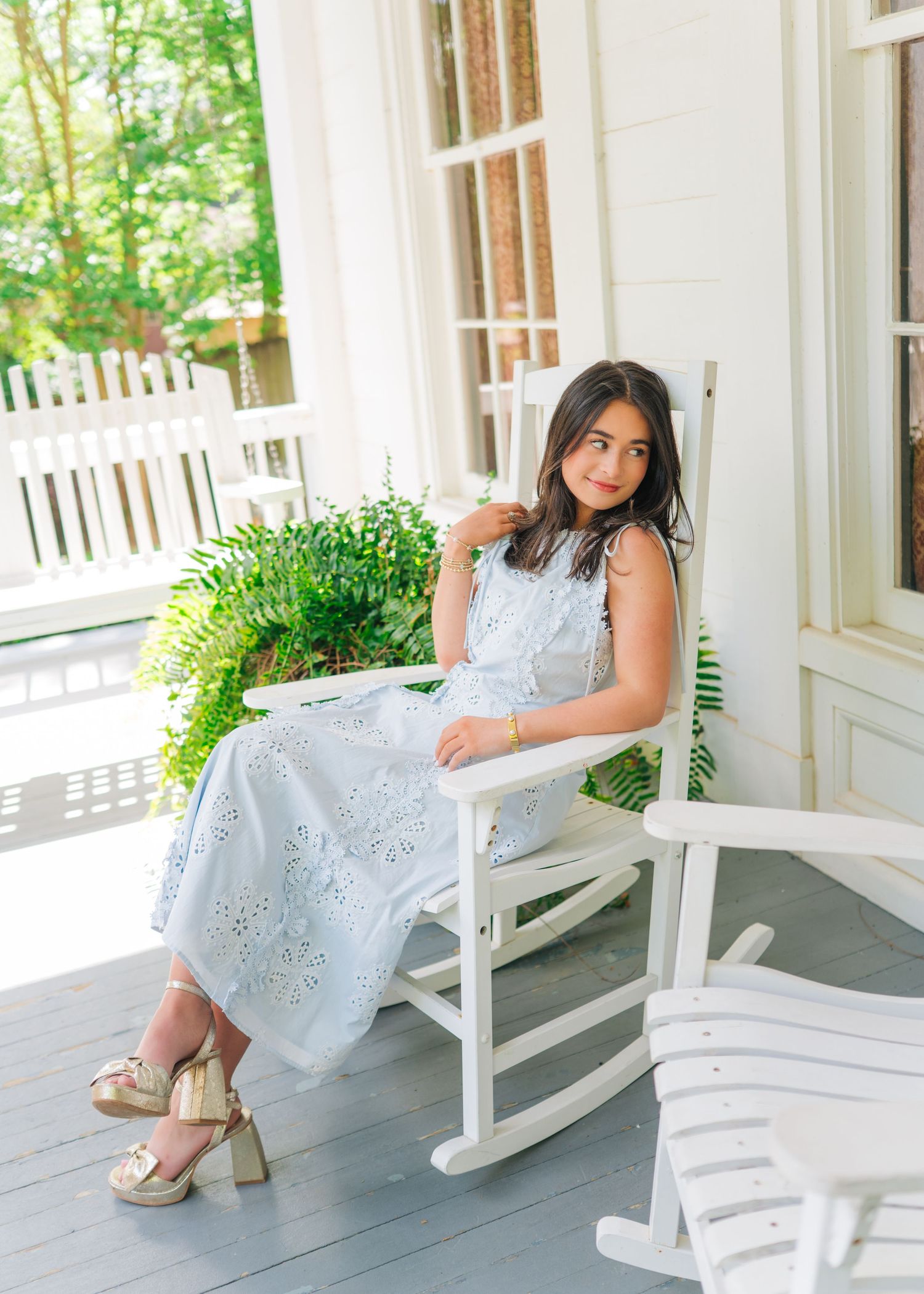 Smiling young woman in white eyelet dress sitting in a rocking chair on a classic white porch.
