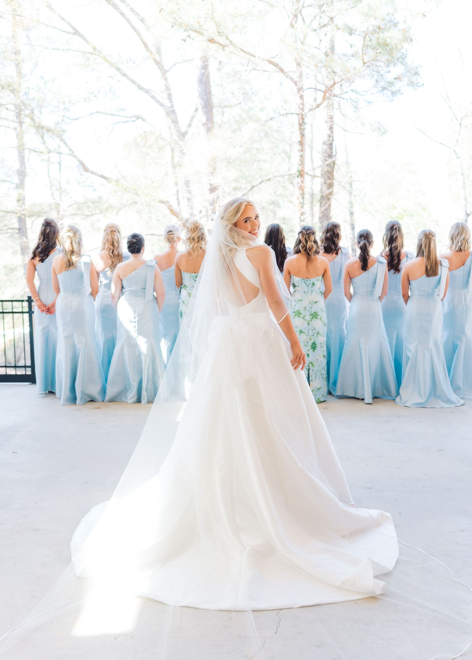 Bride in white gown with veil stands before bridesmaids in light blue dresses outdoors.