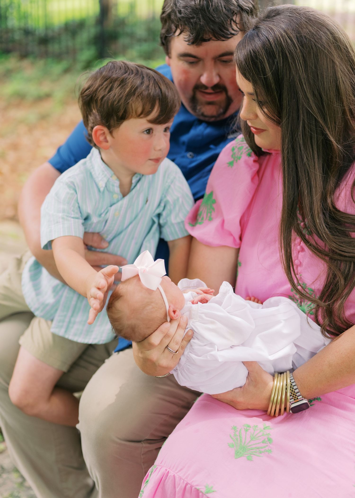 A family of three admires their newborn baby, with a young boy in a blue shirt leaning in curiously outdoors.
