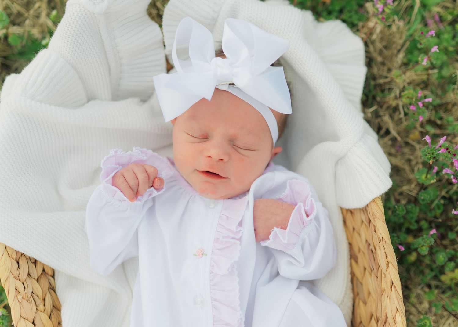 Sleeping newborn baby in white outfit and bow headband resting in a wicker basket with white blanket.