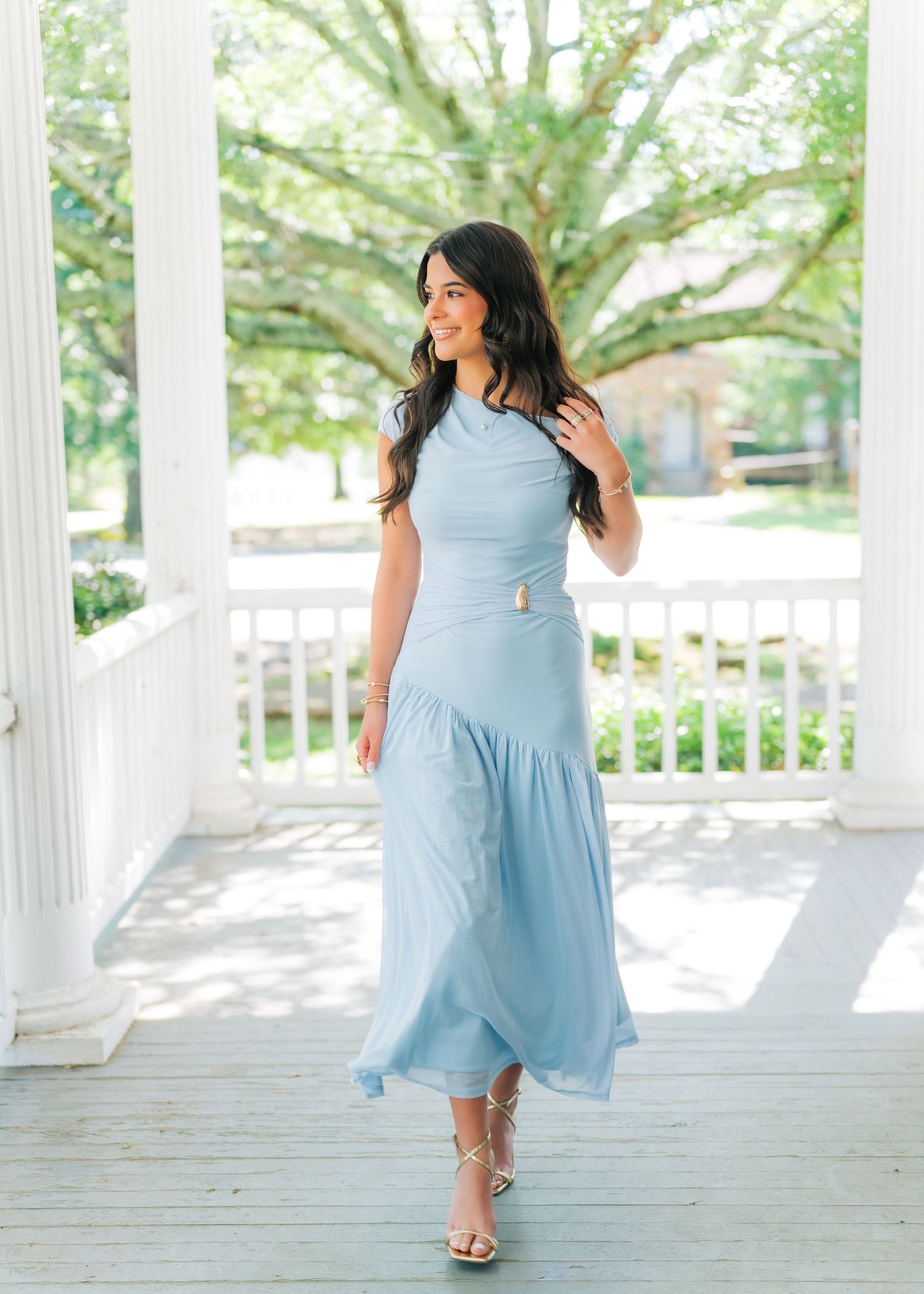 Woman in light blue midi dress with gold accessories poses on white porch.