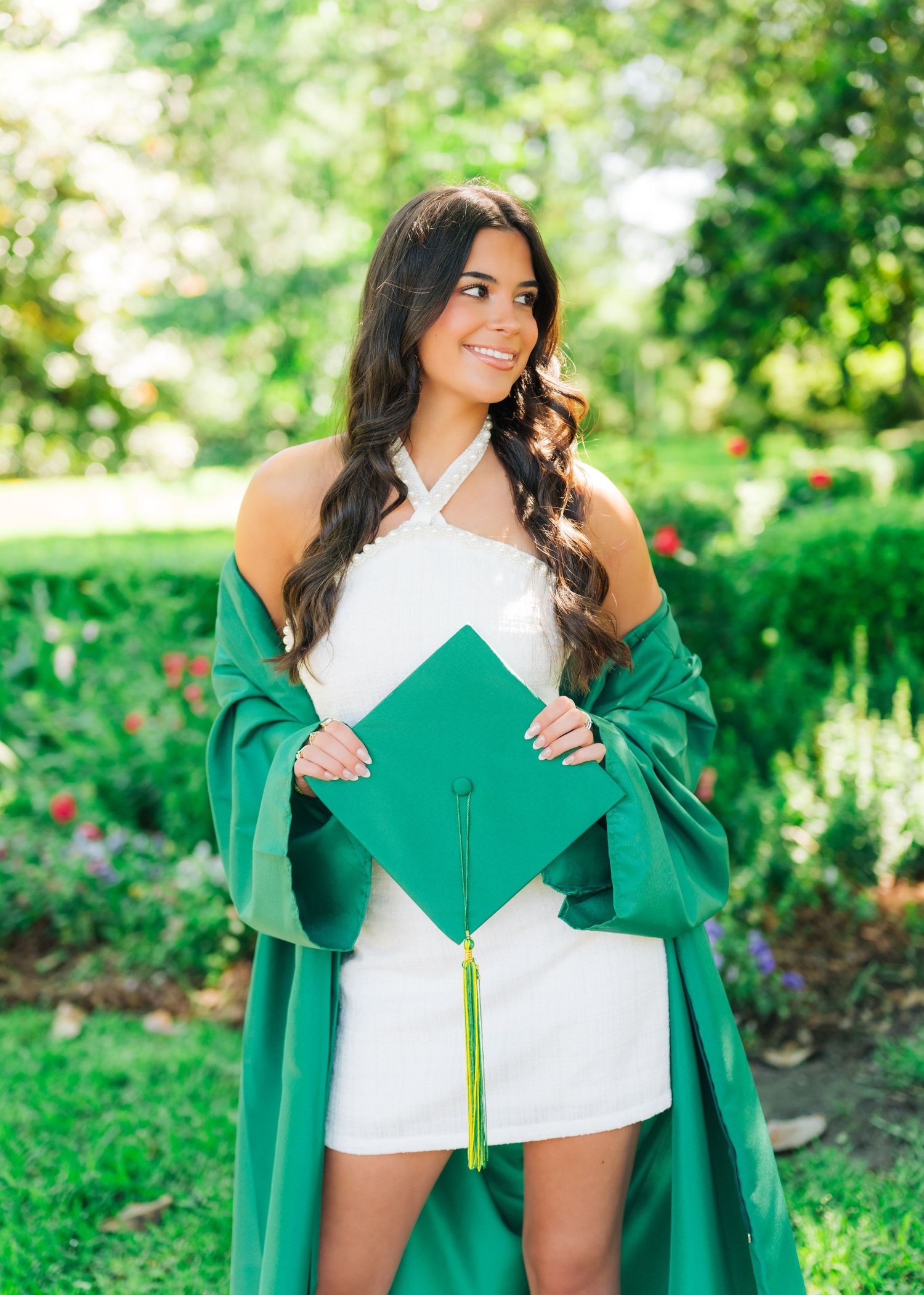 Smiling graduate proudly displaying green mortarboard cap wearing white dress outdoors.