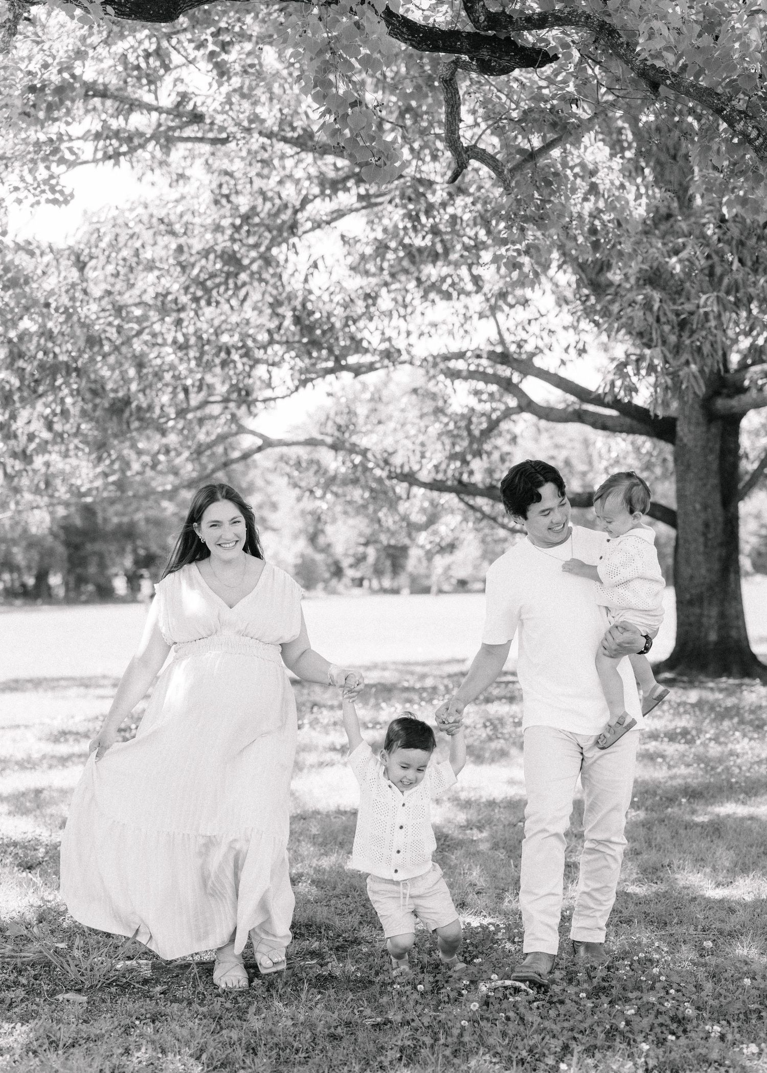 Black and white family photo of pregnant woman, man holding toddler, and young boy walking under a large tree.
