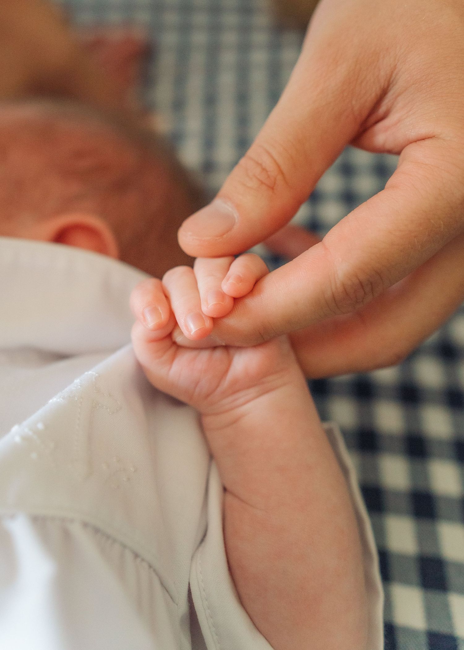 Tender close-up of a newborn baby grasping an adult finger, showcasing tiny delicate hands on gingham fabric.