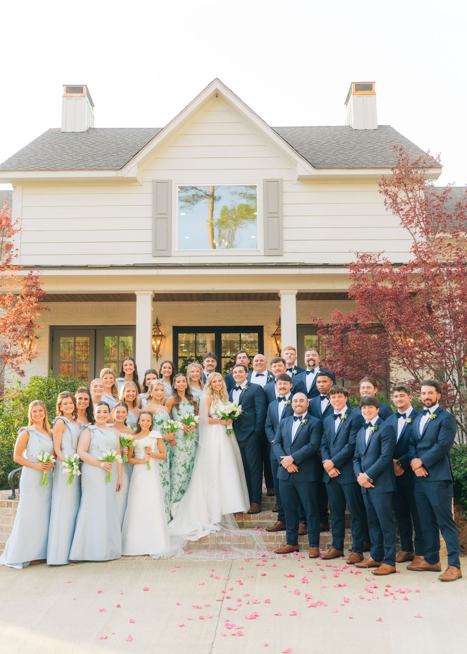 Large wedding party poses in front of white colonial house with bridesmaids in blue and groomsmen in navy.