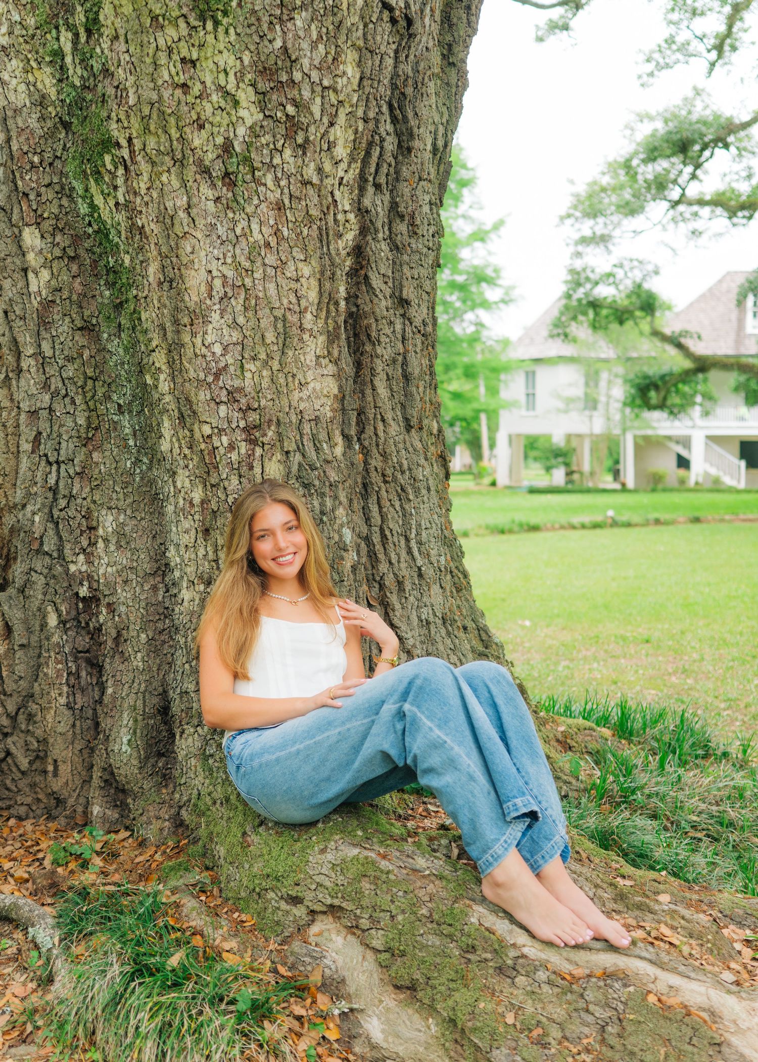 Young woman with long blonde hair sits barefoot against a large tree trunk in a white top and jeans.