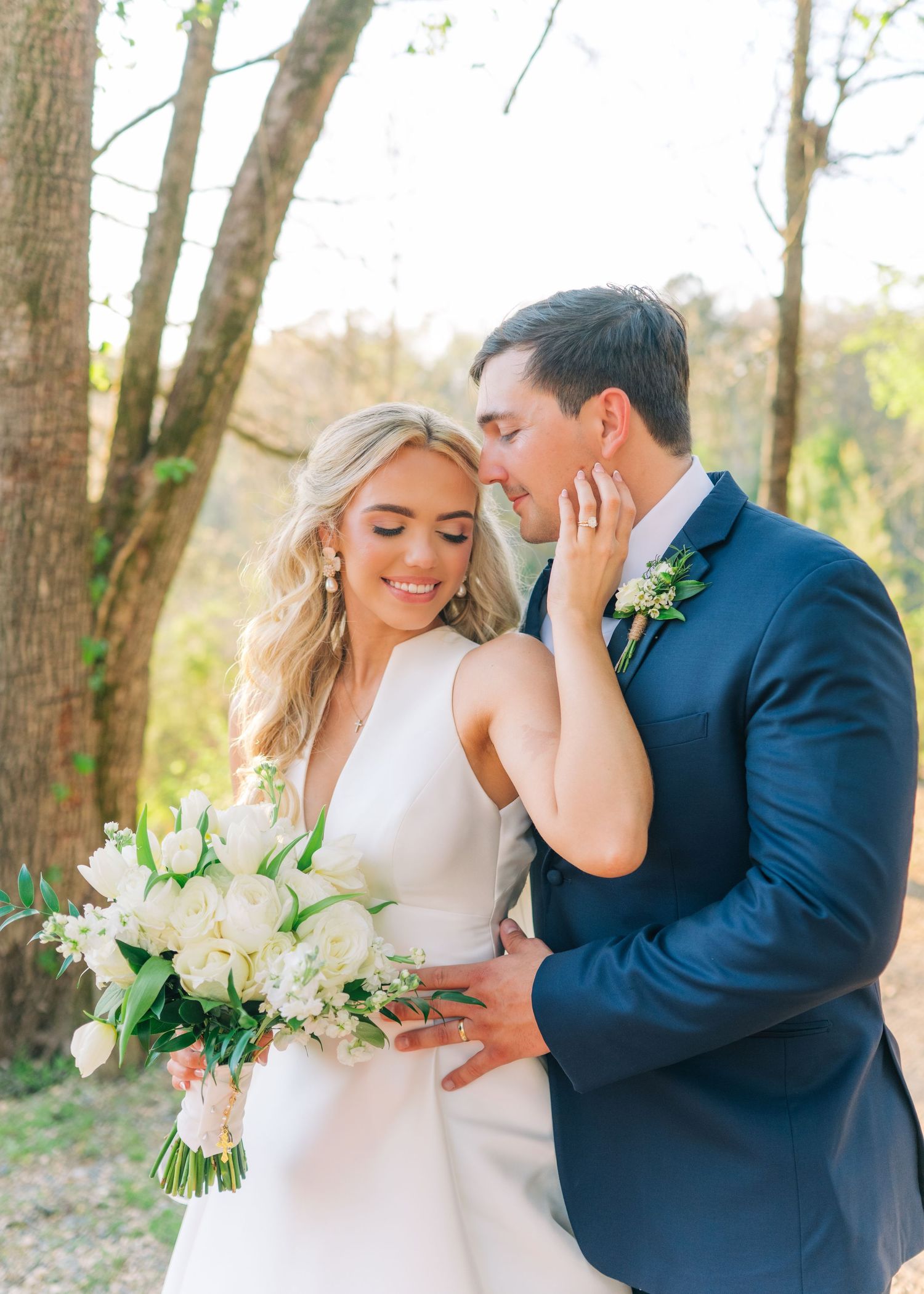 Bride holding white floral bouquet smiles as groom in navy suit whispers to her outdoors among trees.