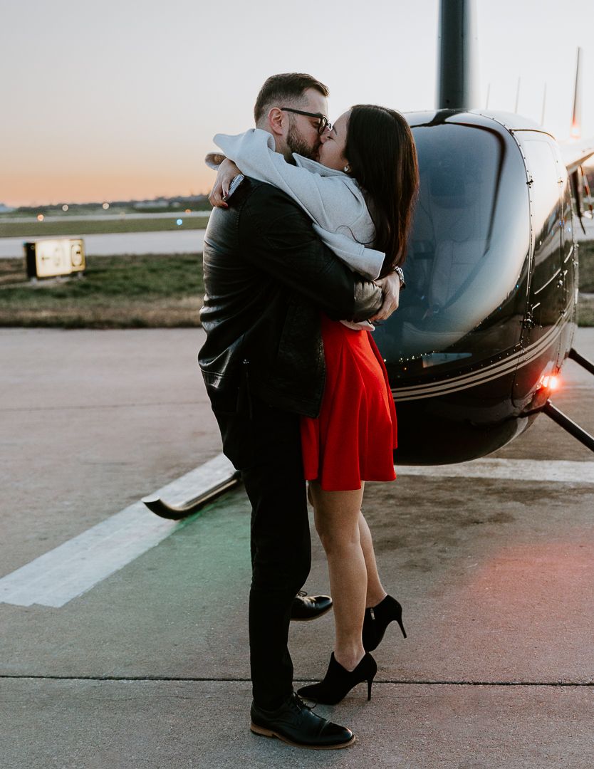 A romantic couple in formal attire shares a tender moment near a black helicopter.