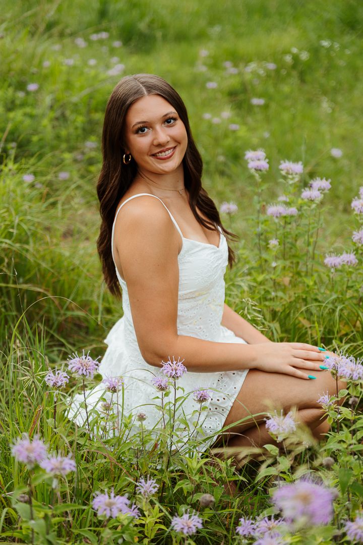Person in white sundress poses among purple wildflowers in a summer meadow.