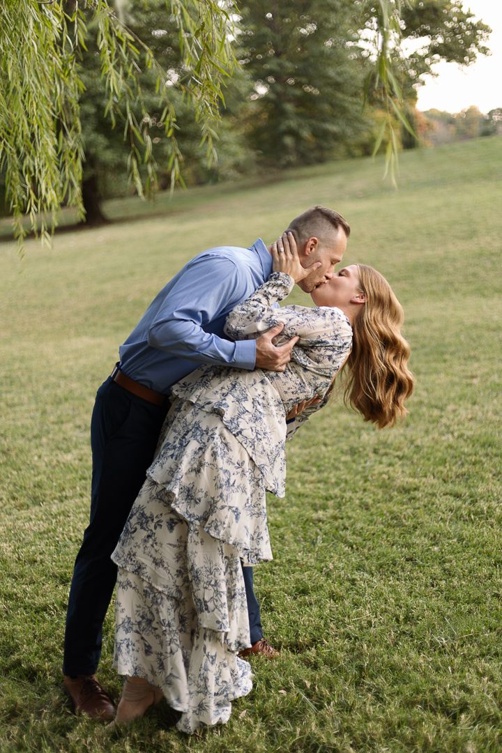 Romantic couple shares a dip kiss on grassy hillside while wearing blue shirt and floral dress at sunset.