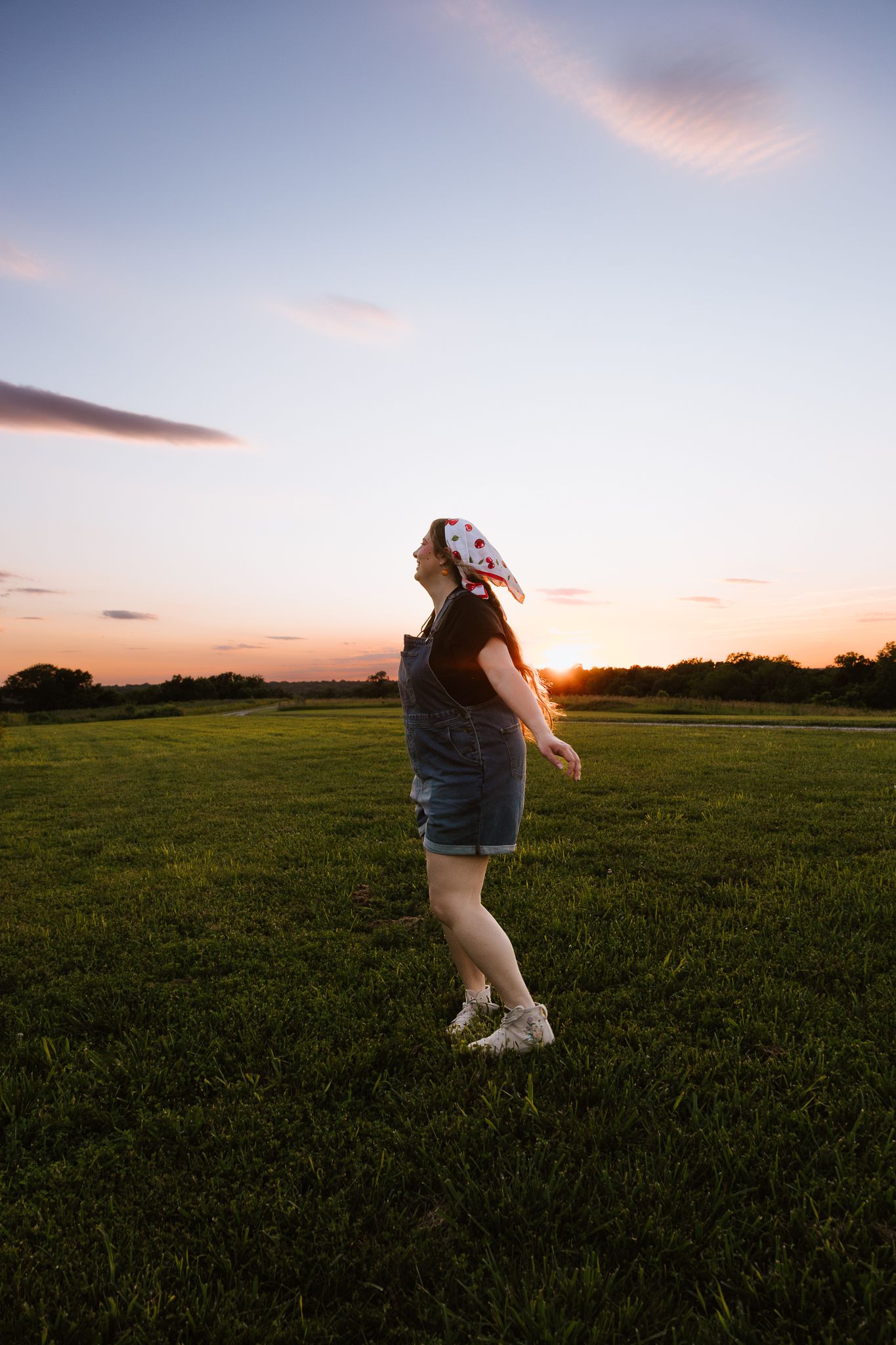 Young woman in denim overalls dancing joyfully in a green field during a vibrant sunset.