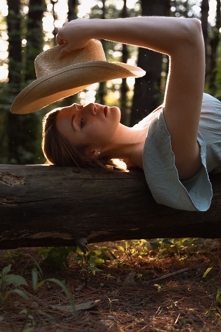 Dreamy sunset portrait of someone reclining on a fallen log in the forest while wearing a straw hat.